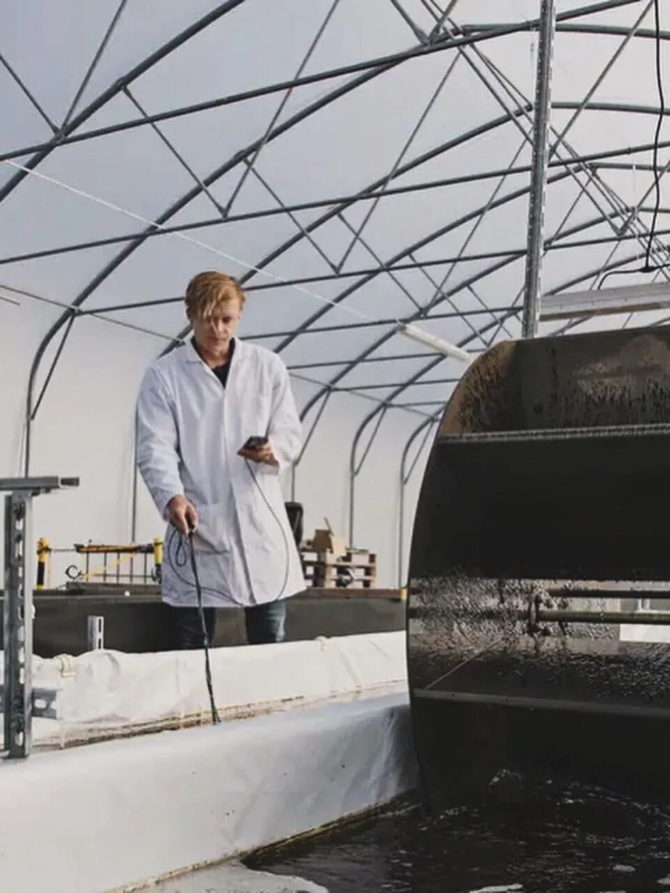 Scientist in a white lab coat conducting experiments in a greenhouse with structural framework overhead, standing by a large black tank or container filled with dark liquid, using a measuring instrument.