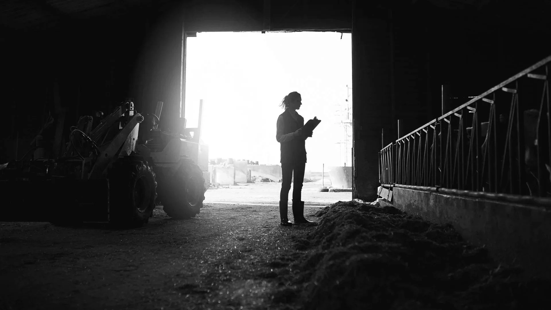 A person standing inside a warehouse, holding a clipboard and looking out towards the light coming in from outside. There are construction or industrial equipment to the left and a metal railing on the right.
