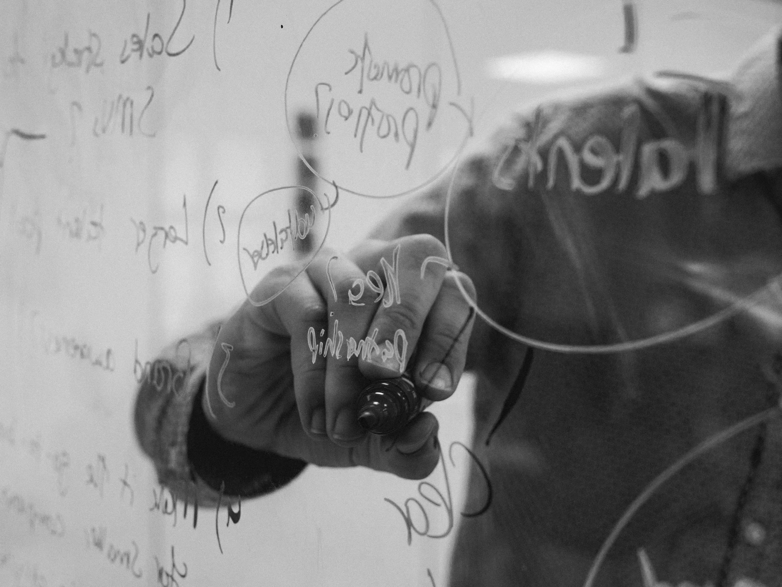 A person writing on a transparent surface with a dark marker, with handwritten notes and diagrams visible on the surface.