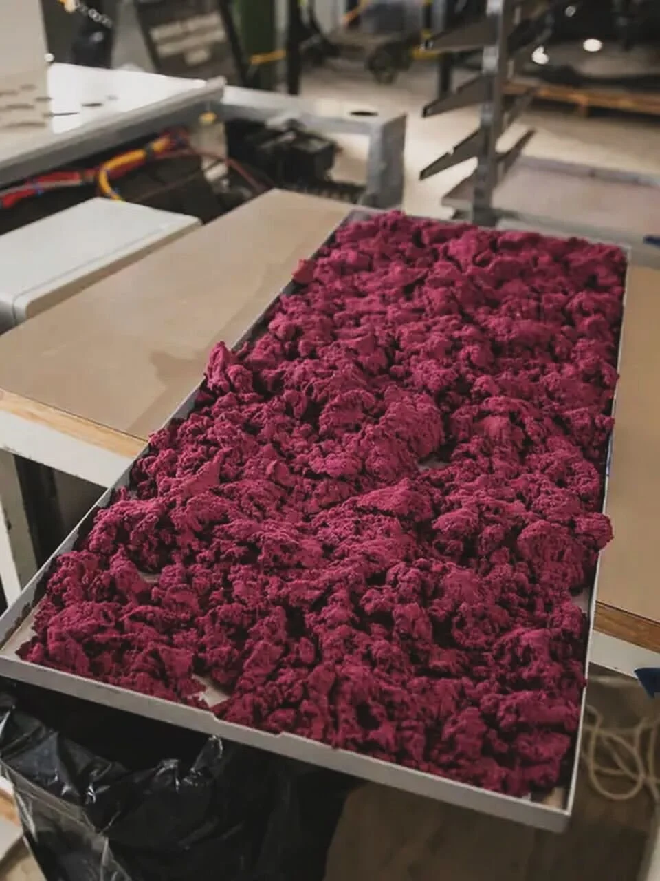 A tray filled with red, textured, sponge-like material, possibly foam or synthetic fabric, placed on a workbench in a workshop or factory setting.