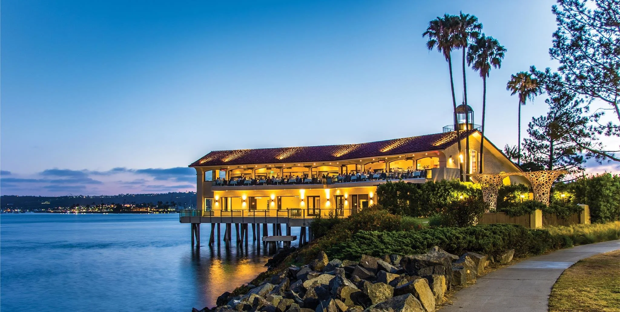 Restaurant over water with outdoor dining area, palm trees, and stone decorations at dusk.