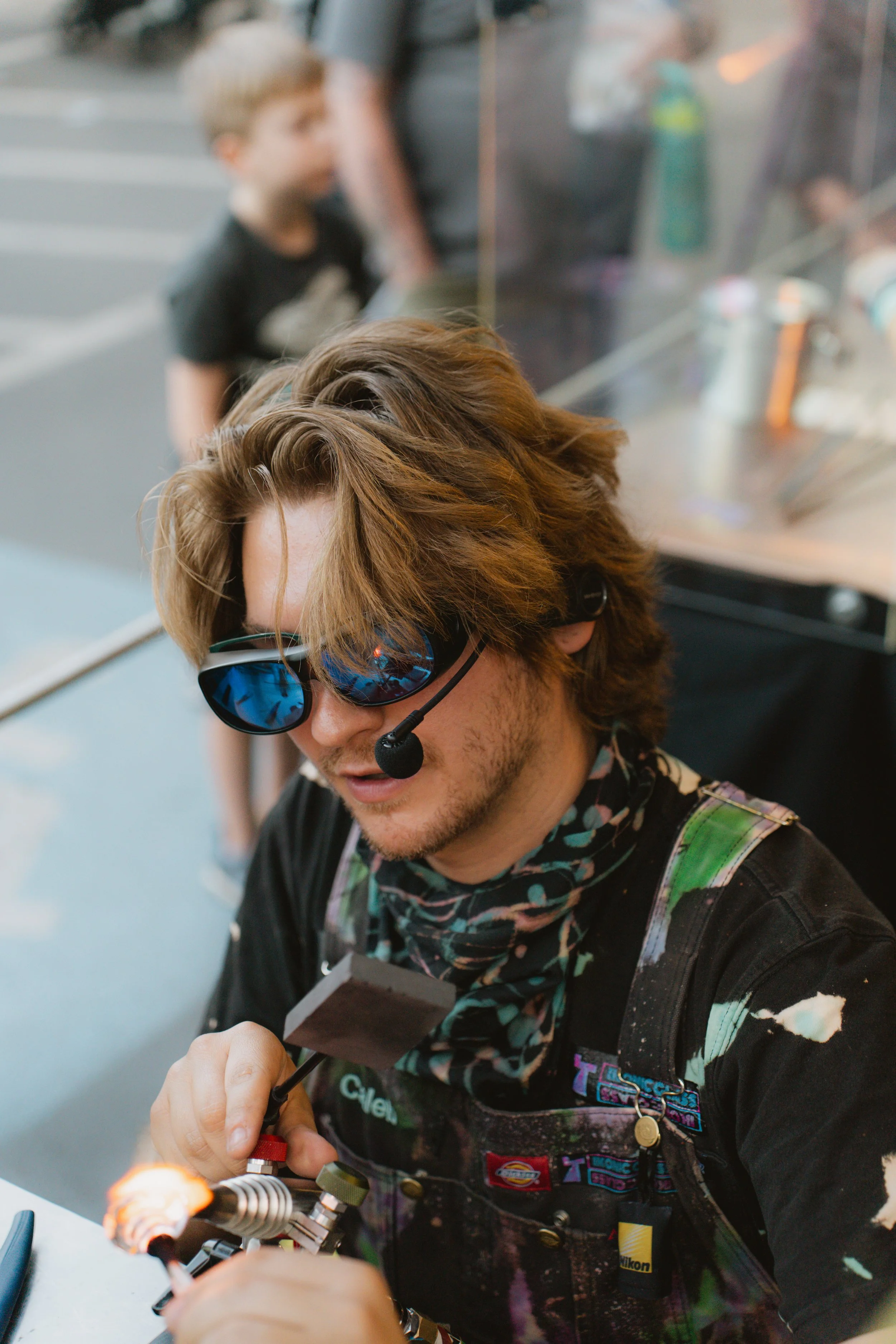 Man with curly hair, sunglasses, and black shirt blowing fire using a blowtorch in a workshop.
