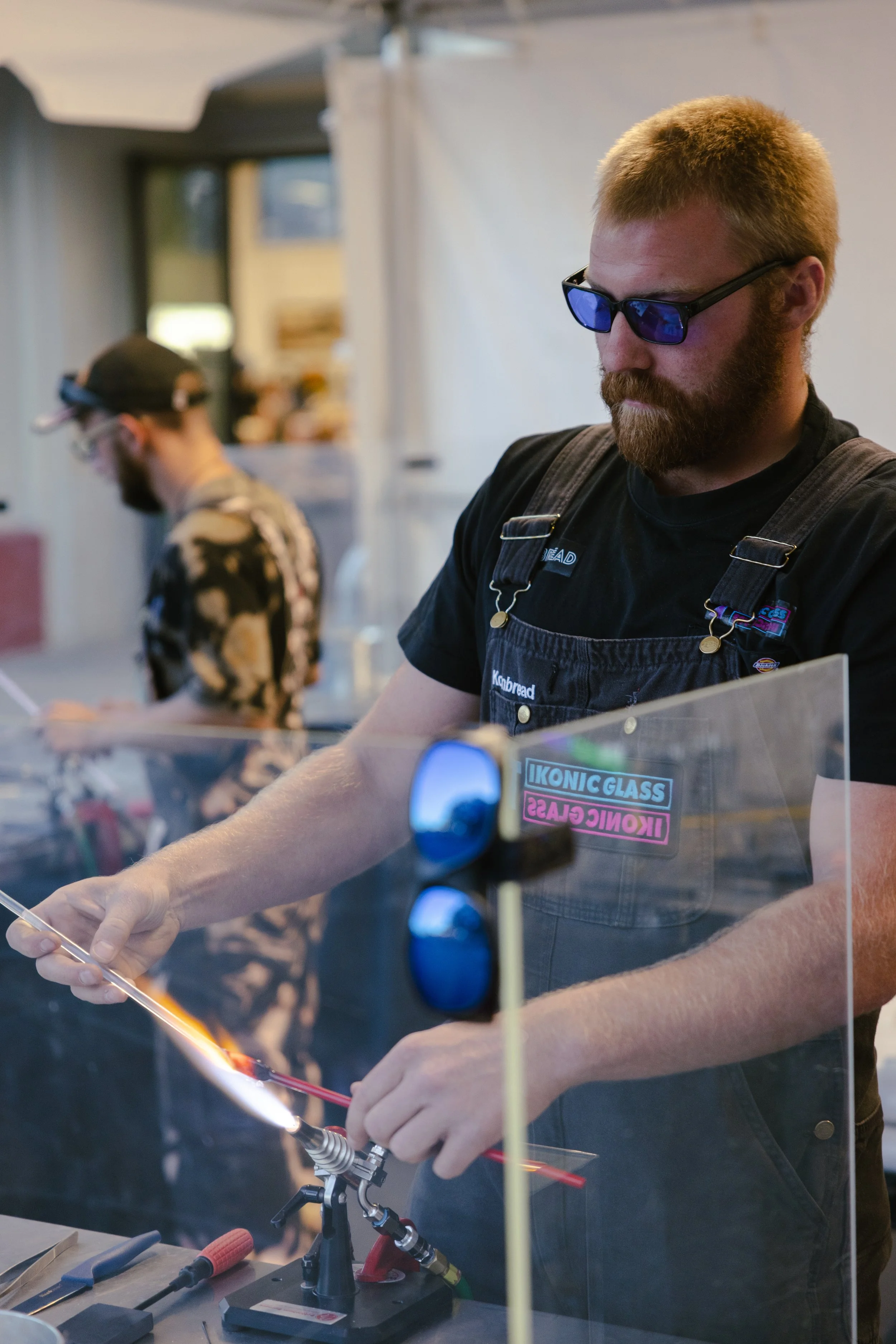 A man with red hair and beard wearing sunglasses and a striped shirt, holding a spoon and preparing to stir a purple liquid.