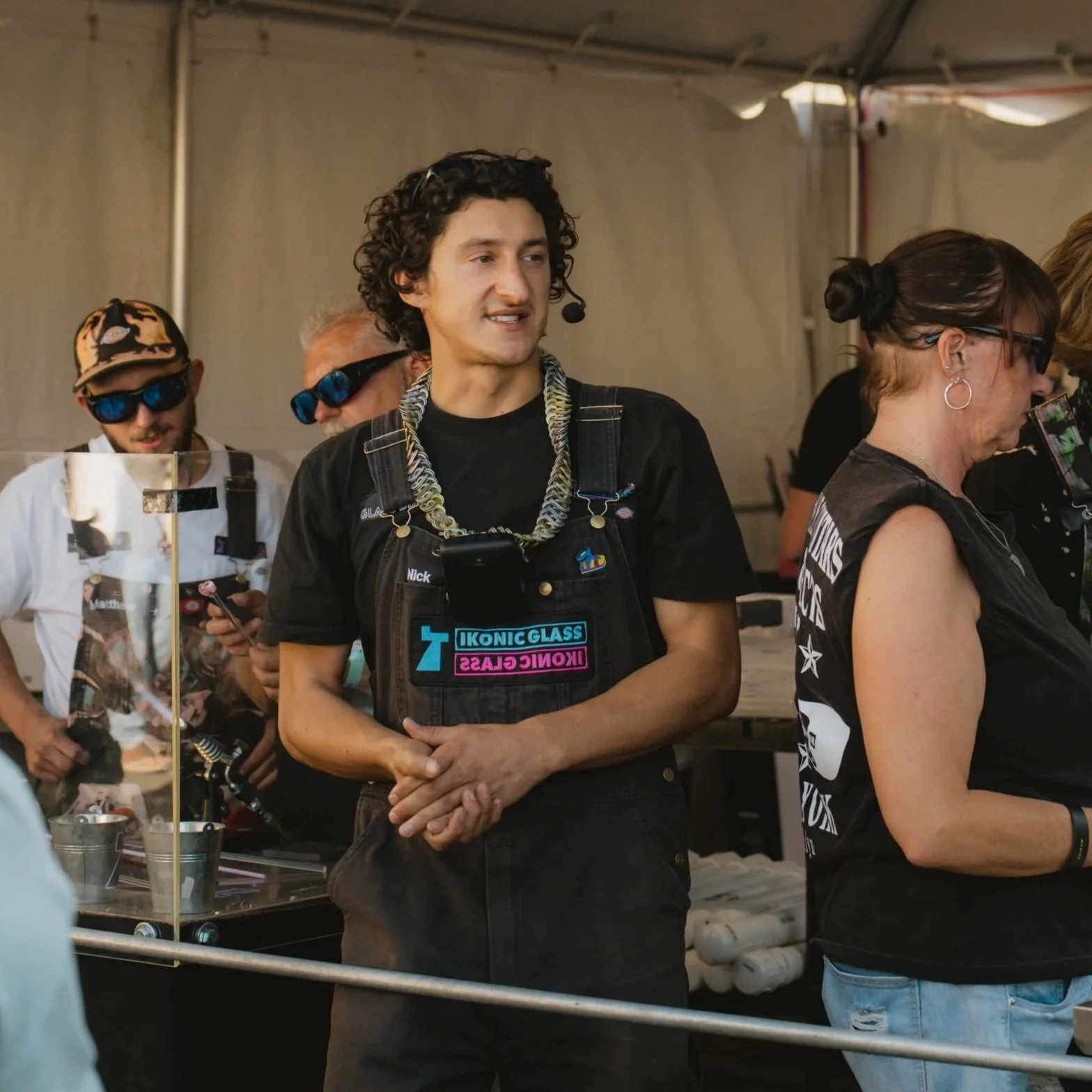 A man with curly dark hair wearing a light purple polo shirt is pointing at a display at an outdoor event or market stall, with other people and tents visible in the background.