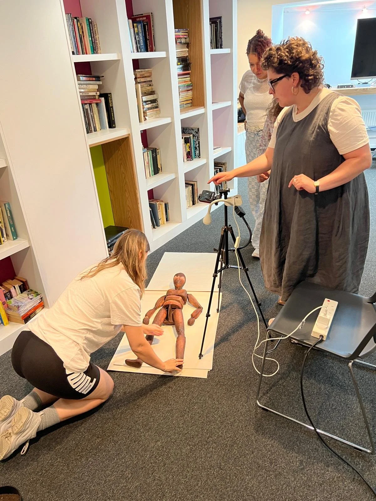 Three people in a studio, are setting up a scene in a room with bookshelves. One person kneels to adjust a large wooden mannequin on paper, while another holds a camera on a tripod, and the third watches. 