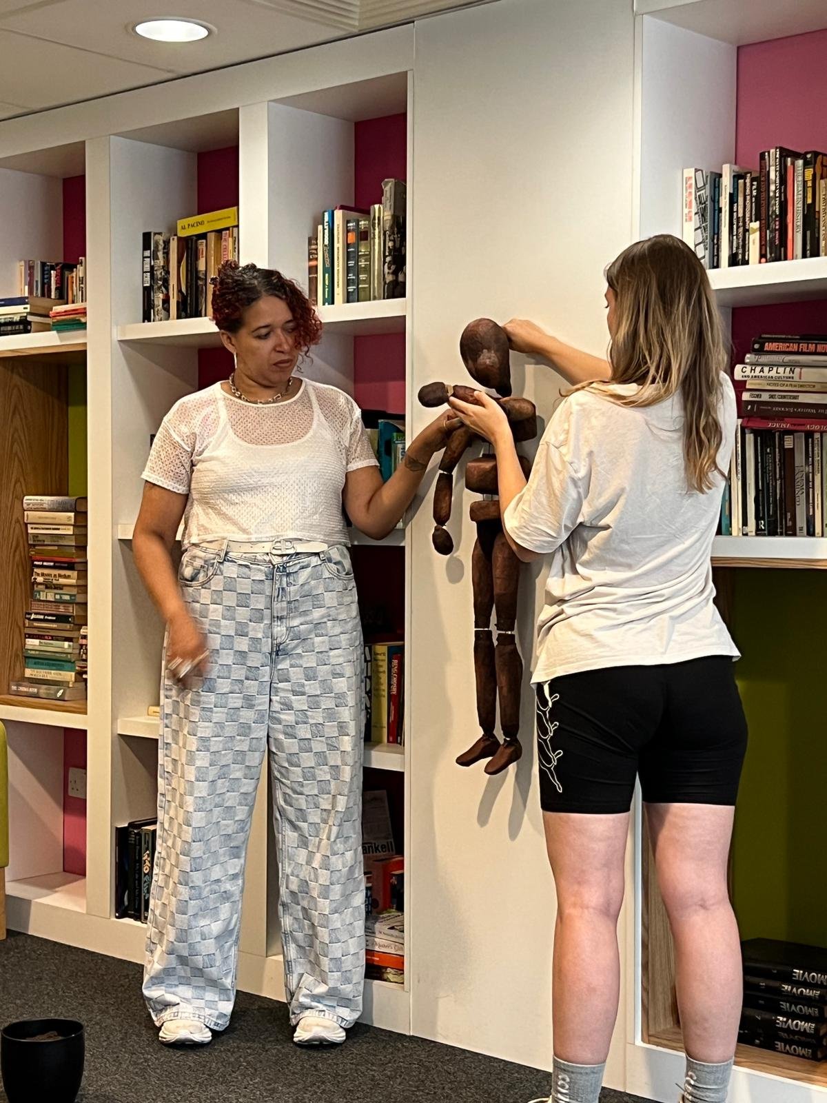 Two people interact with a wooden puppet in a library setting. One person holds the puppet while the other assists. Books line the shelves in the background.