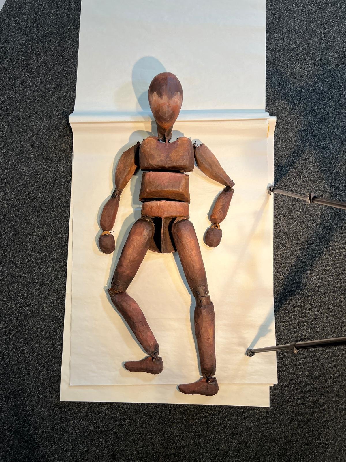 A wooden figure is positioned on a sheet of paper. A black camera tripod is in the corner of the photo.