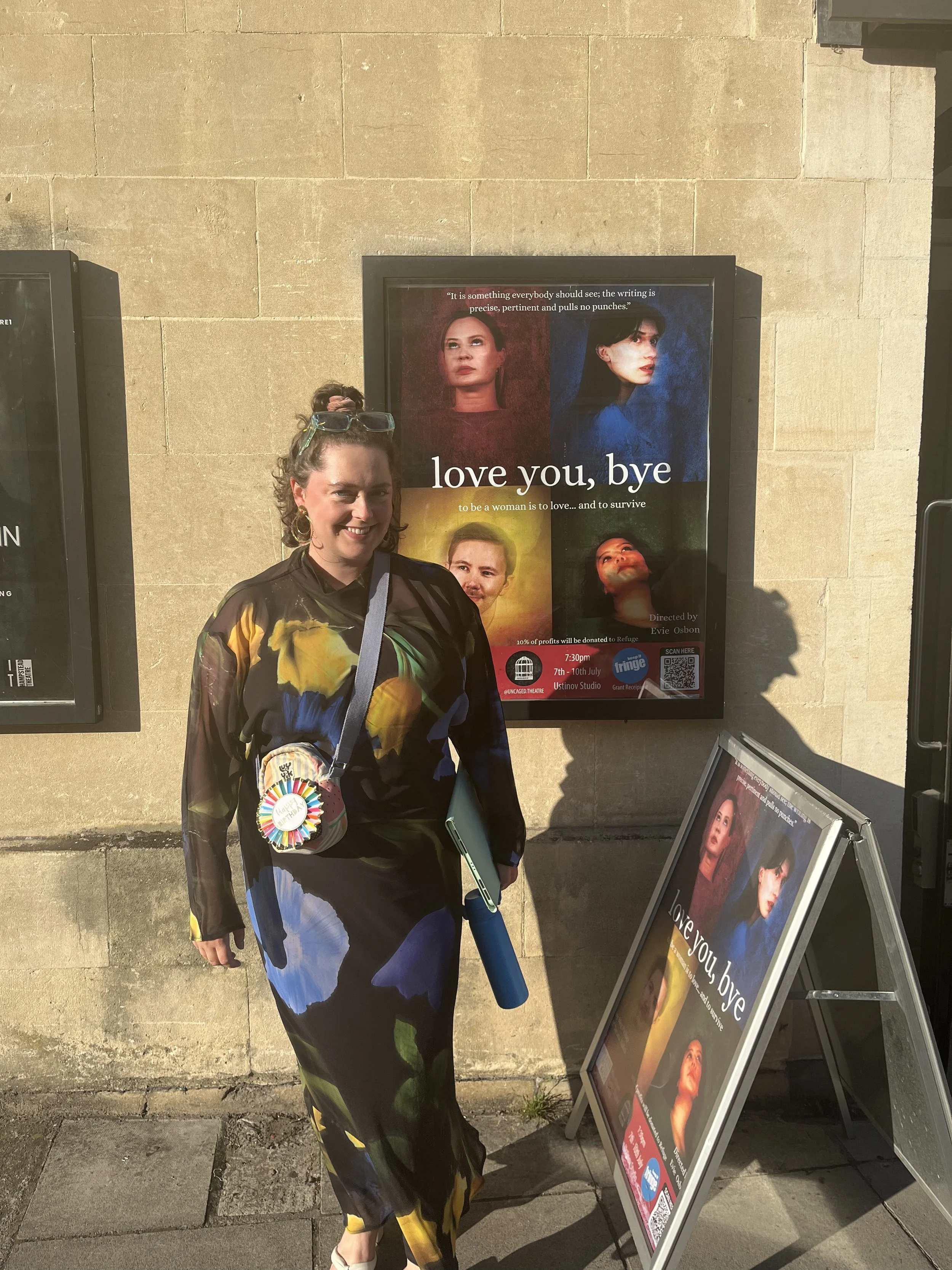 A person stands outside in bright sunlight against a stone wall, next to posters for a production titled “love you, bye.” They are wearing a long, flowing dress with bold abstract patterns in yellow, blue, green, and black, accessorized with a colorf