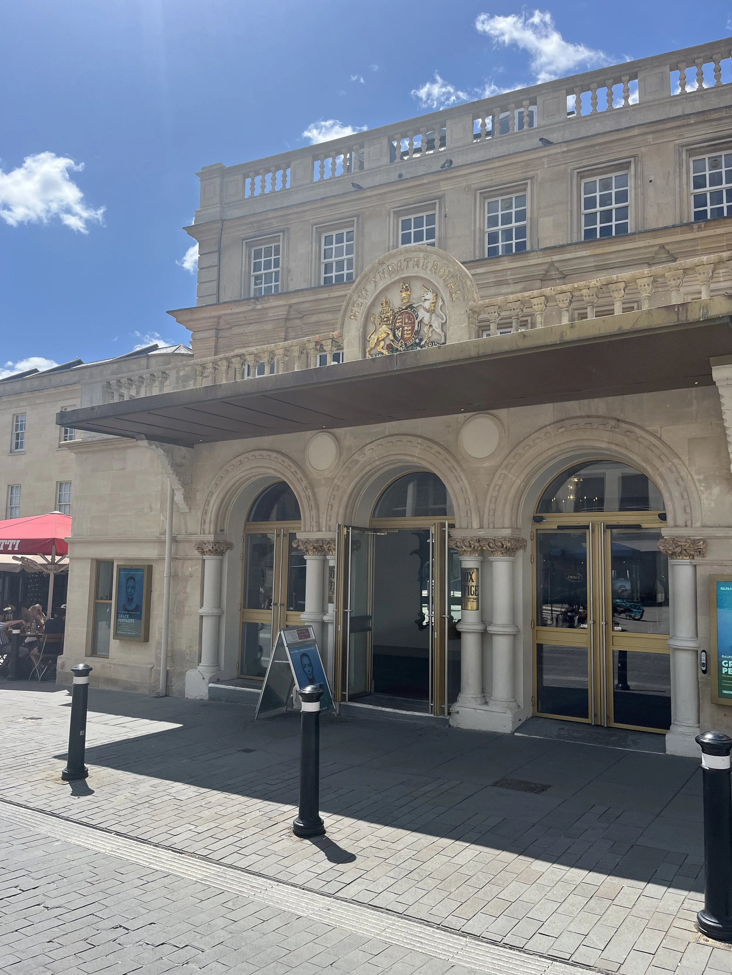 The front entrance of the Theatre Royal building, featuring three arched doorways with decorative stone columns and gold-framed glass doors. Above the entrance is a crest set into the stonework. The scene is brightly lit by sunlight, with blue sky ov