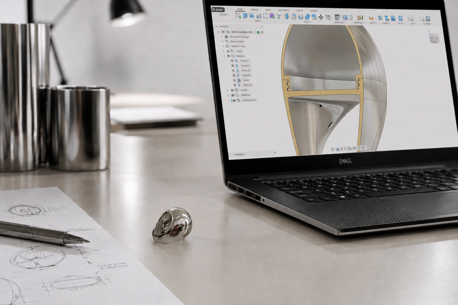 Design software on a Dell laptop screen showing a 3D model of a mechanical part. The workspace includes toolbars, layers, and design options. The desk has technical drawings, a metallic pen, a shiny ring, and metallic containers, with a desk lamp in the background.