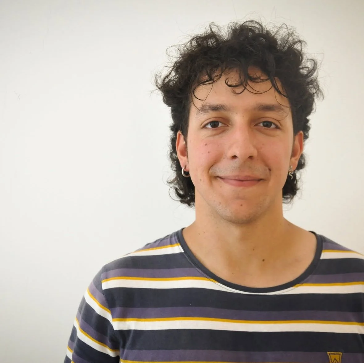 A young man with curly dark hair, light skin, and earrings, wearing a striped t-shirt, smiling at the camera against a plain white background.
