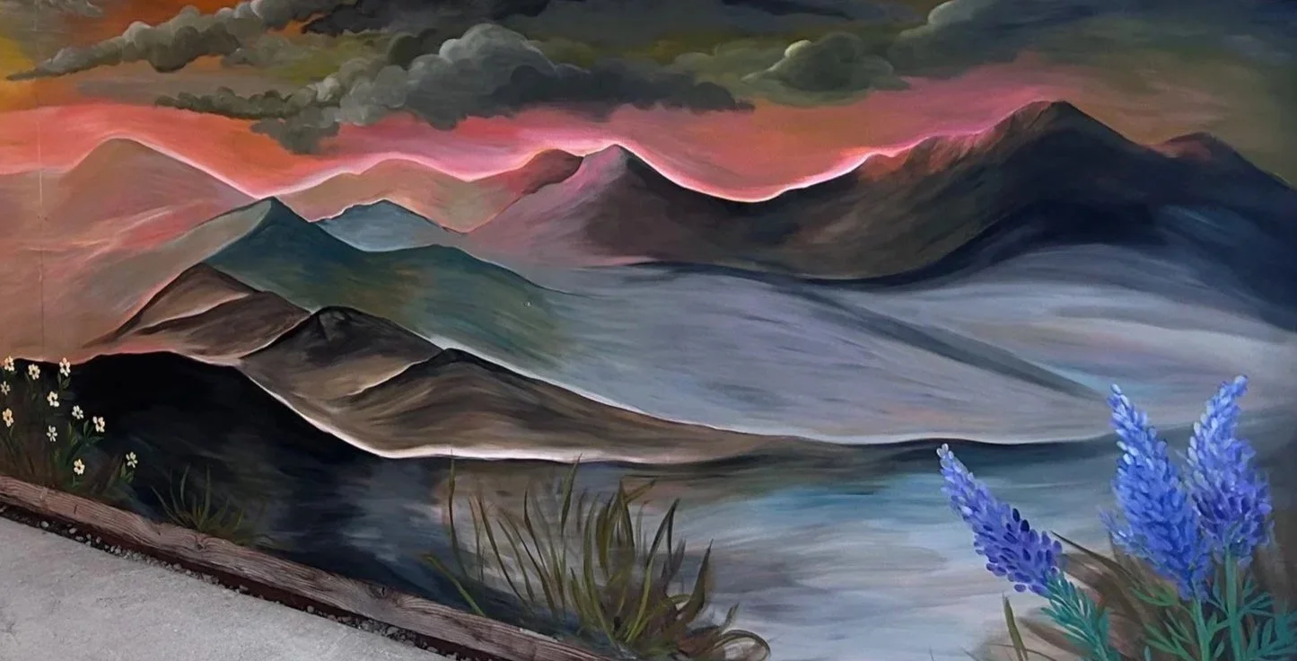 A colorful landscape painting of mountains, a lake, and plants. The mountains are in shades of blue, green, and purple, with some peaks crowned by dark clouds. The sky is a blend of pink and orange, suggesting sunset or sunrise. In the foreground, there are white daisies on the left and tall purple flowers on the right, with grass and foliage surrounding them.