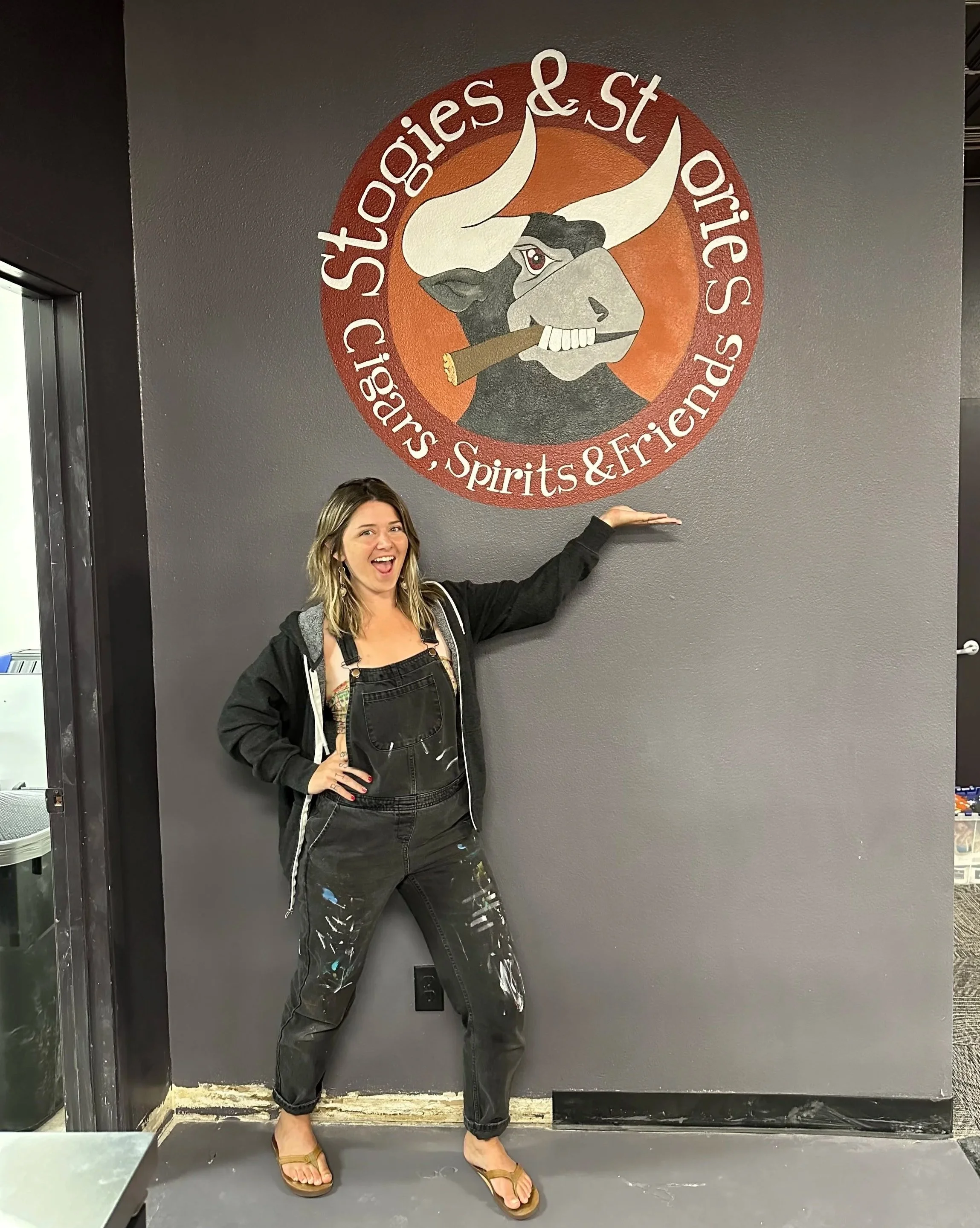 A woman in black pants, a black hoodie, and tan sandals is standing in front of a wall with a large circular logo that features a cartoon bull holding a cigar and the text 'Stoogies & Stogies, Spirits & Friends' around the circle.
