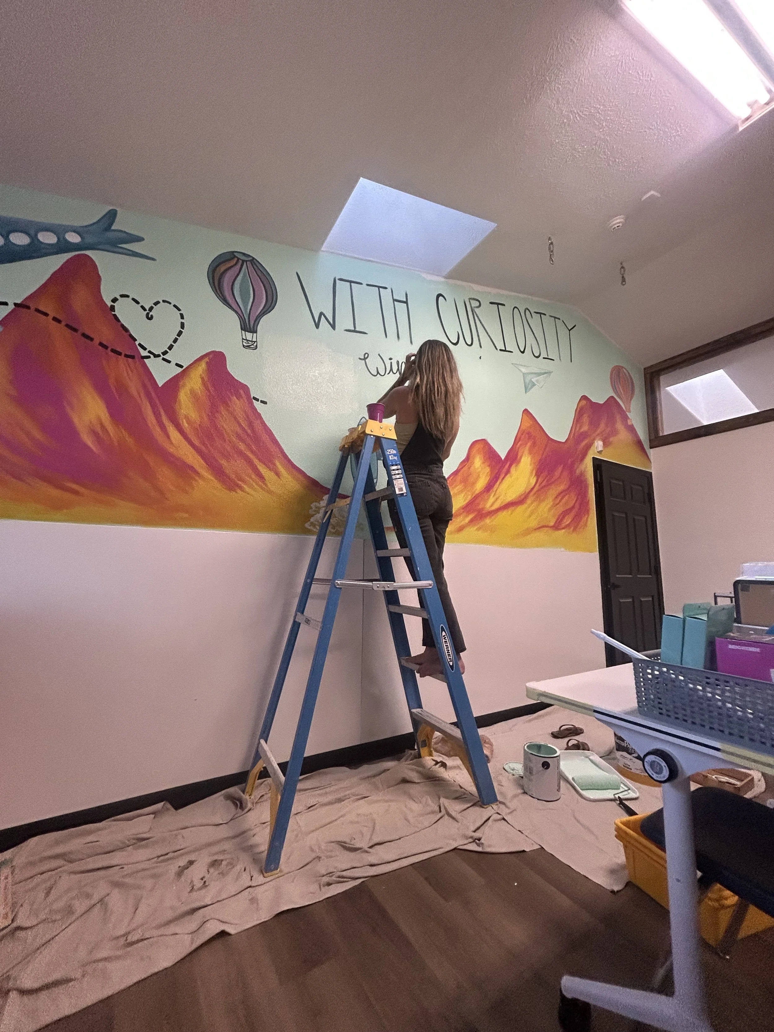 A person is painting a colorful mural on a wall in a room. They are standing on a ladder, working on the artwork, which features mountains, hot air balloons, and the phrase 'WITH CURIOSITY' written on the wall. The room has a drop ceiling, a window, and art supplies on the floor and nearby surfaces.