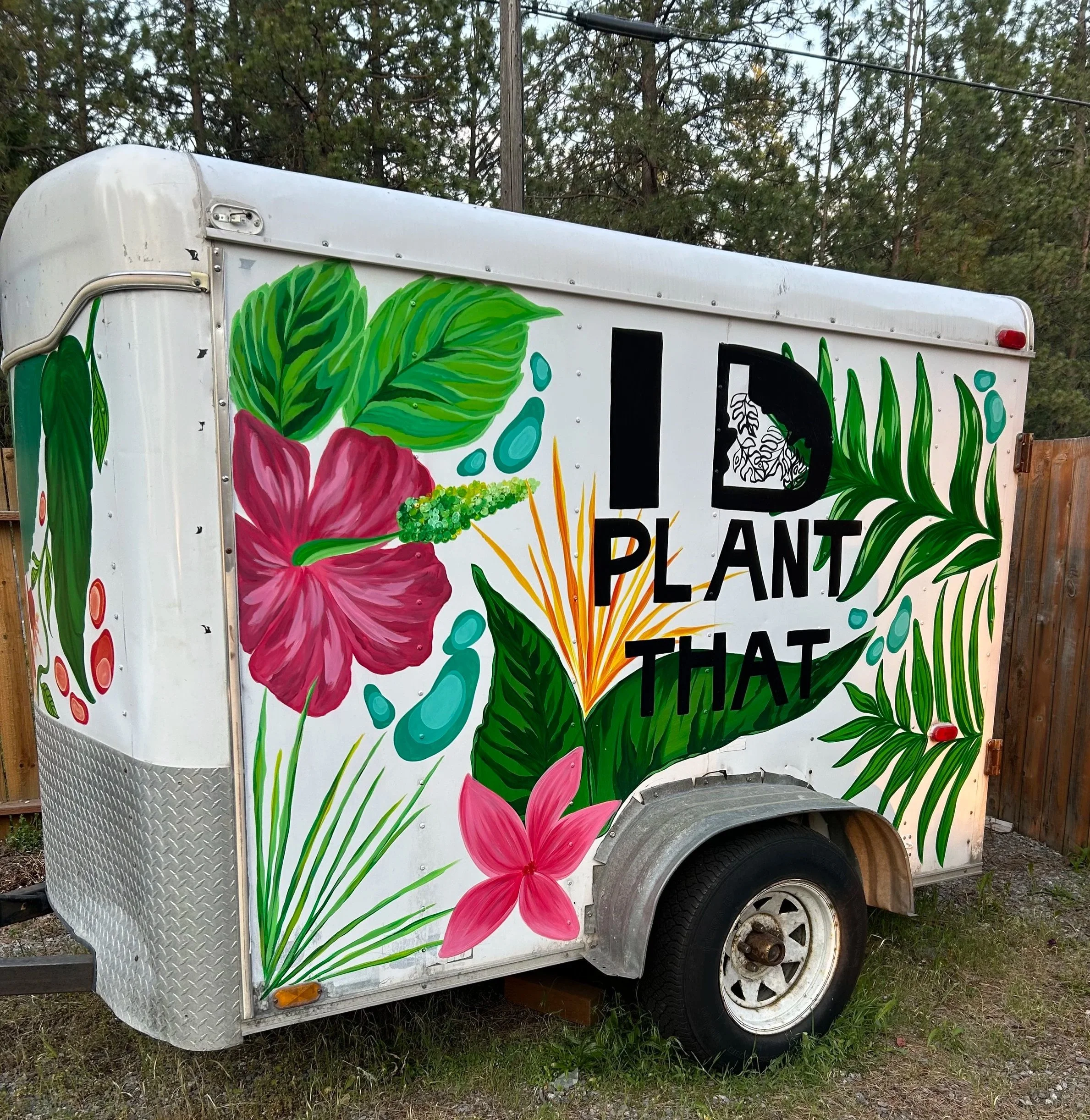 A paint-covered food truck decorated with colorful tropical flower and leaf designs, parked outdoors on grass with trees and utility lines in the background.