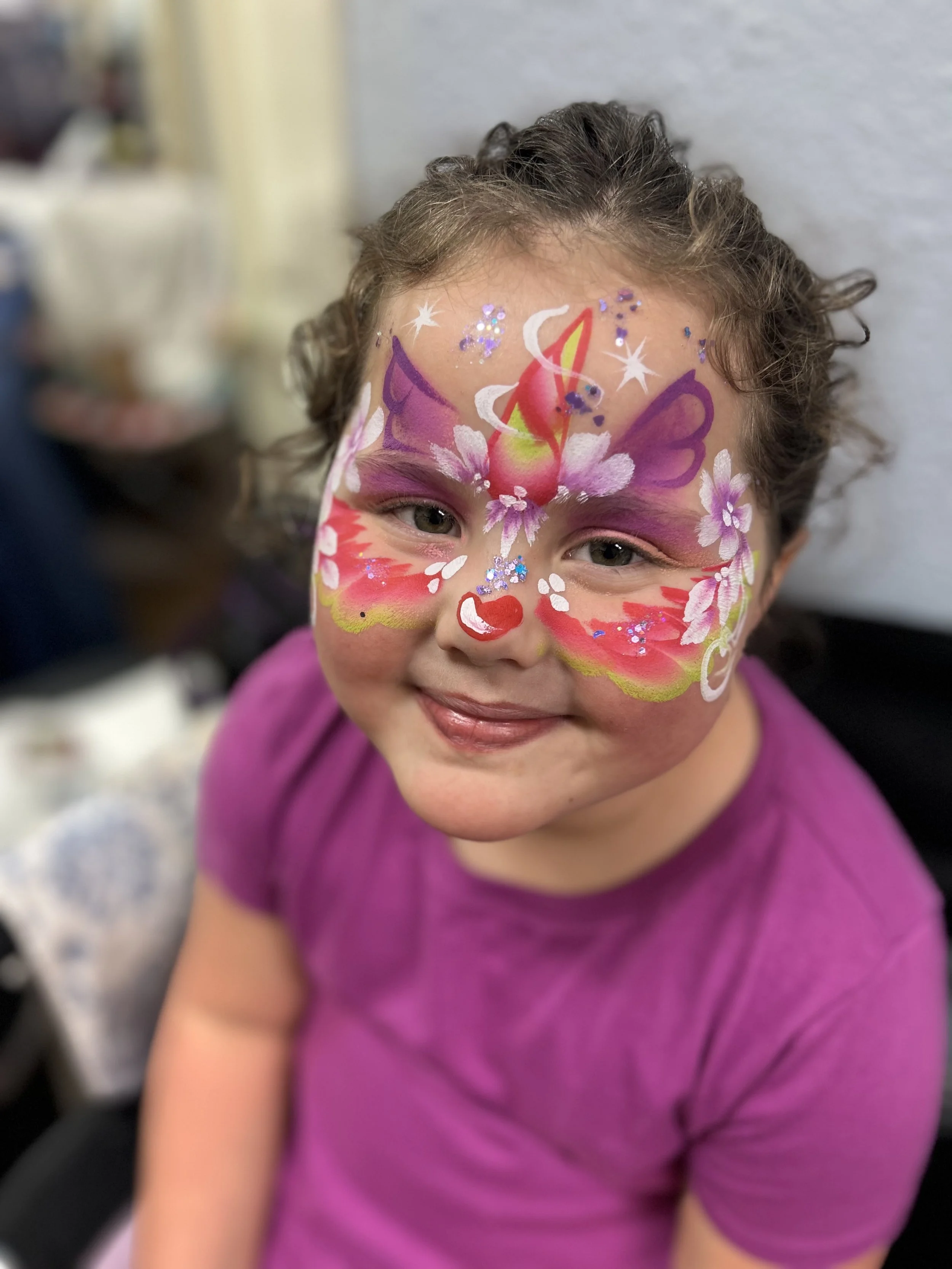 Young girl with long brown curly hair, wearing a purple shirt, has detailed face paint of a butterfly and a colorful unicorn on her face, with glitter embellishments.