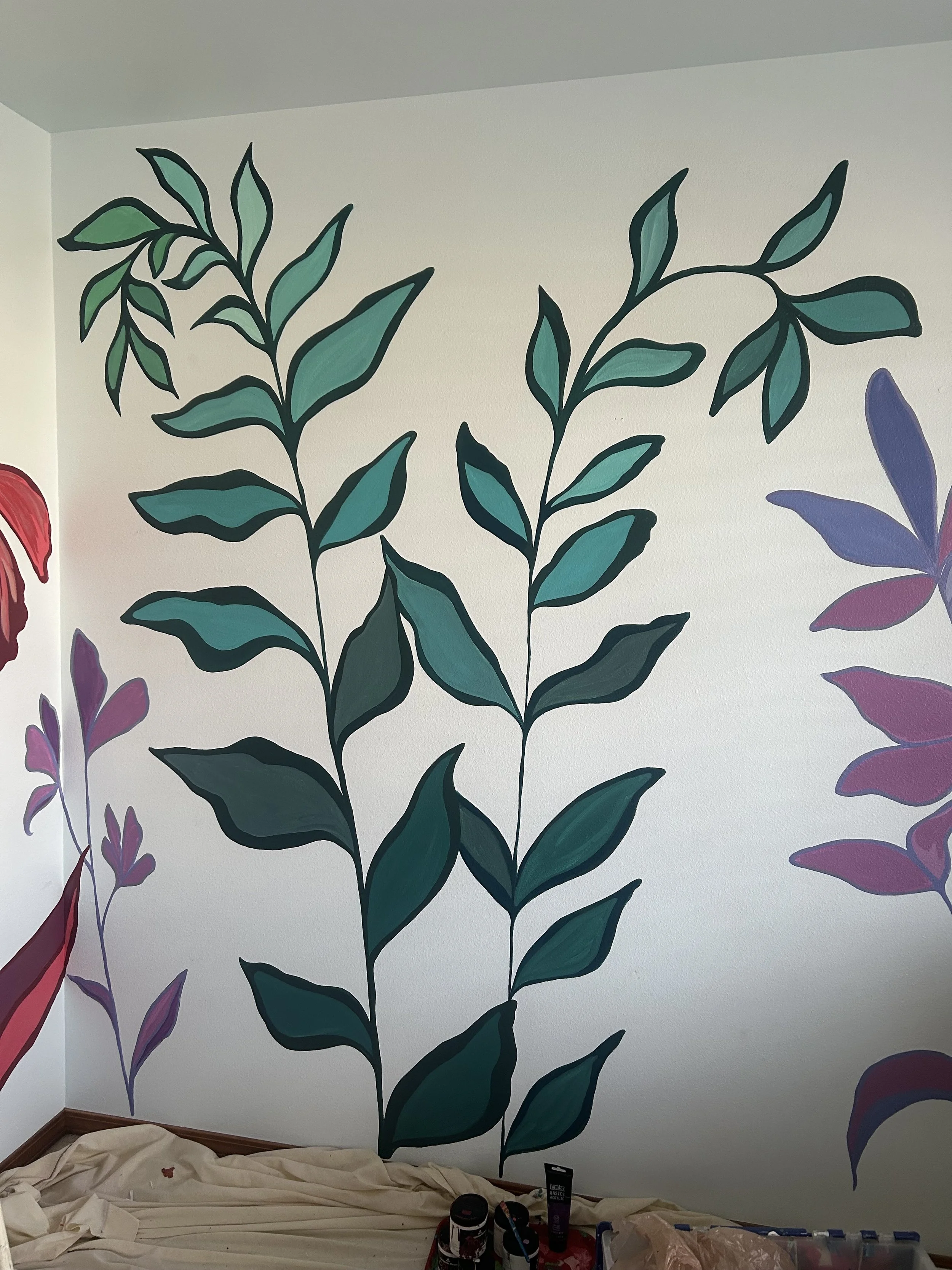 Interior wall with large painted green and purple leafy plant design and some painting supplies and cloth on the floor.