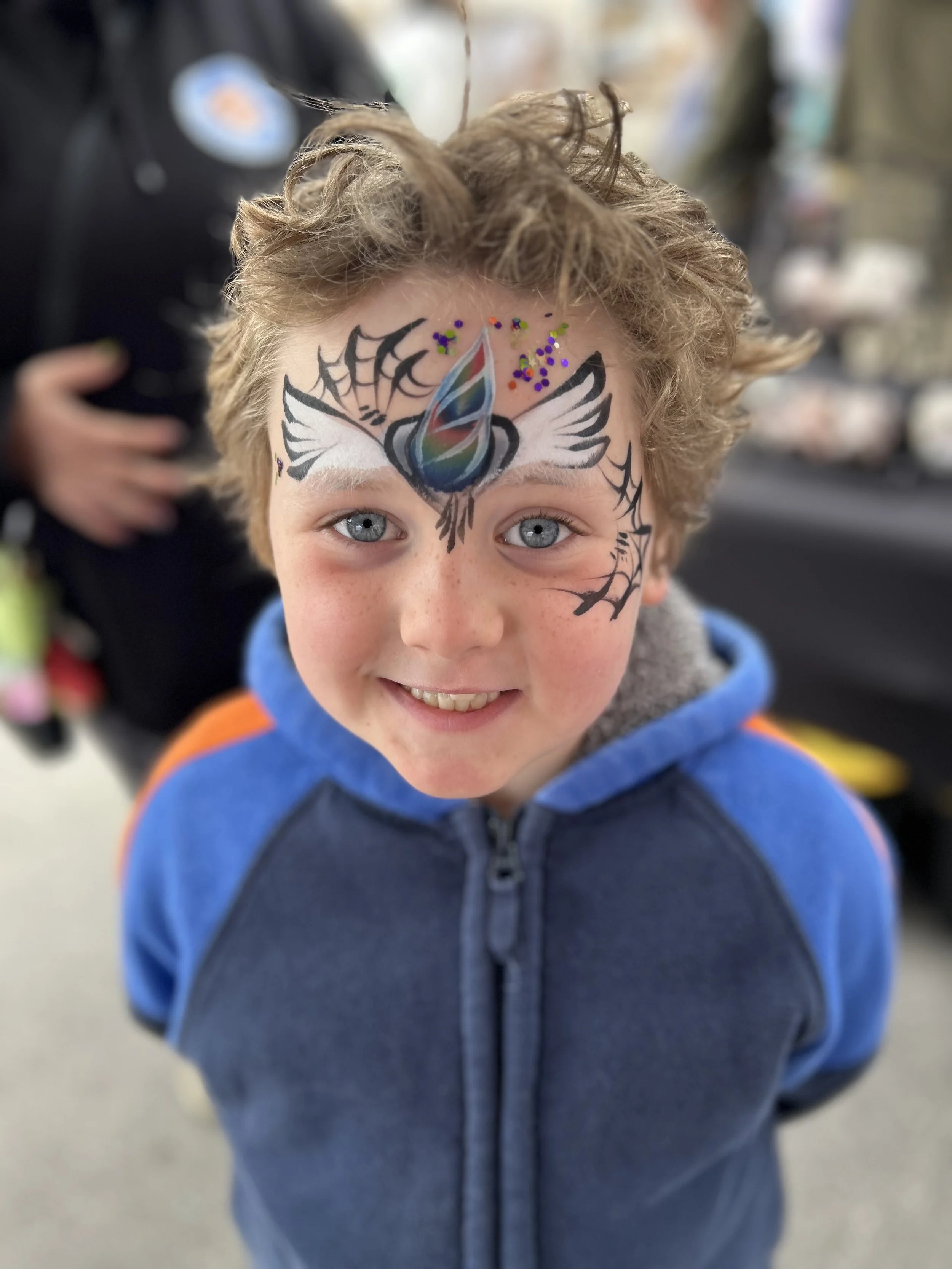 A young boy with curly blond hair and blue eyes smiling at the camera, with face paint of a colorful butterfly and spider web designs on his forehead.