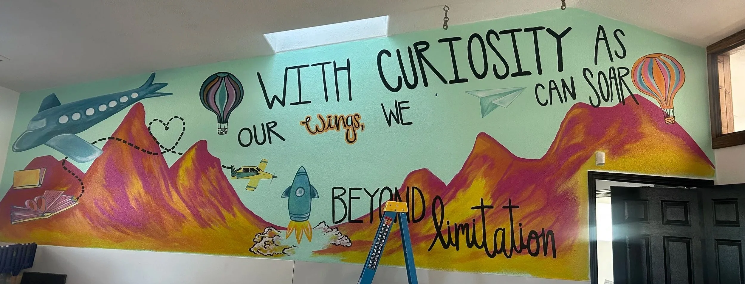 Colorful mural with mountains, flying planes, hot air balloons, a paper airplane, and a rocket. The mural features the quote 'With curiosity as our wings, we can soar beyond limitation.'