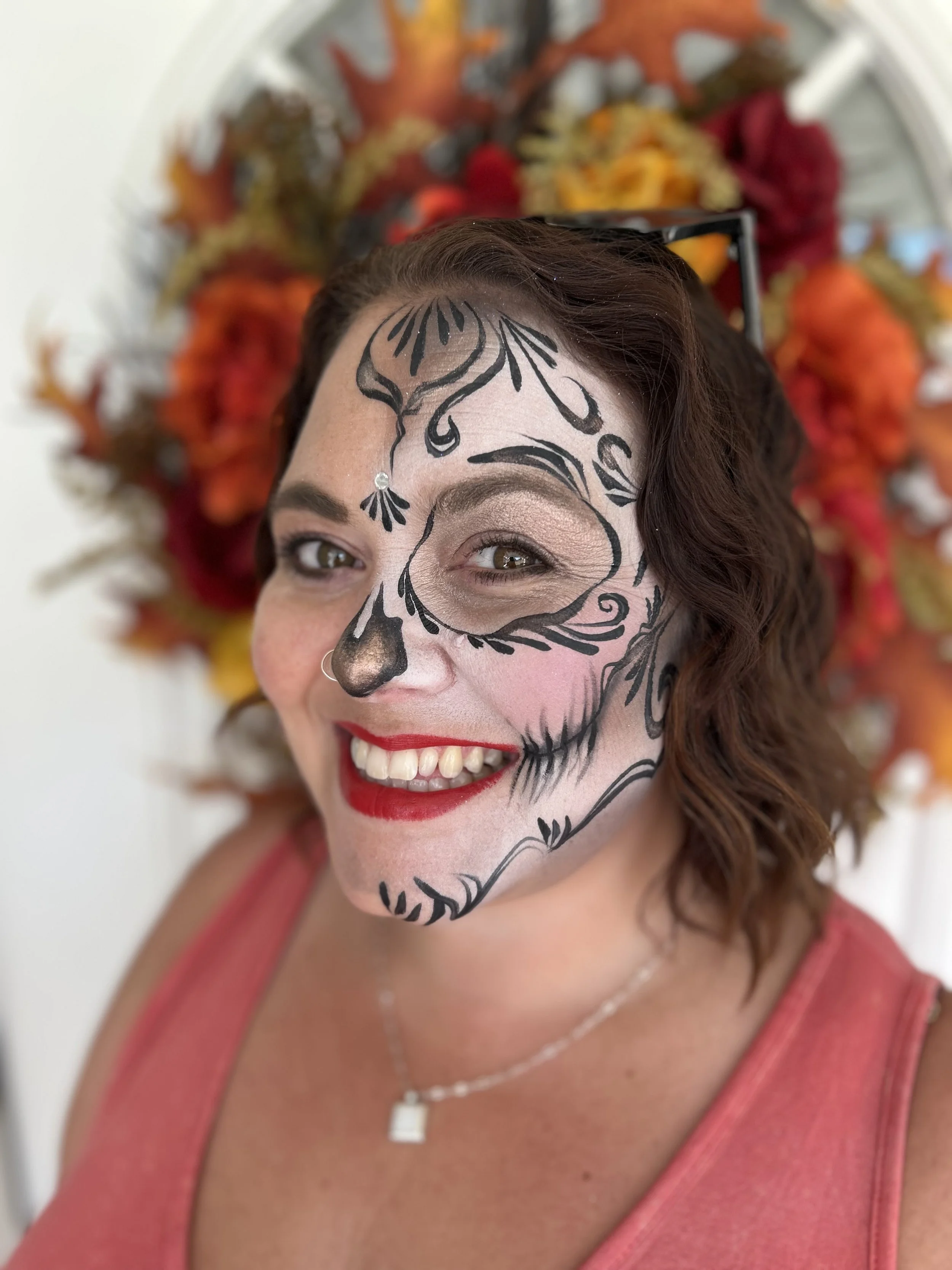 Woman with face painted as a sugar skull, smiling, wearing glasses on her head, red lipstick, pink tank top, and a necklace, with a colorful floral backdrop.