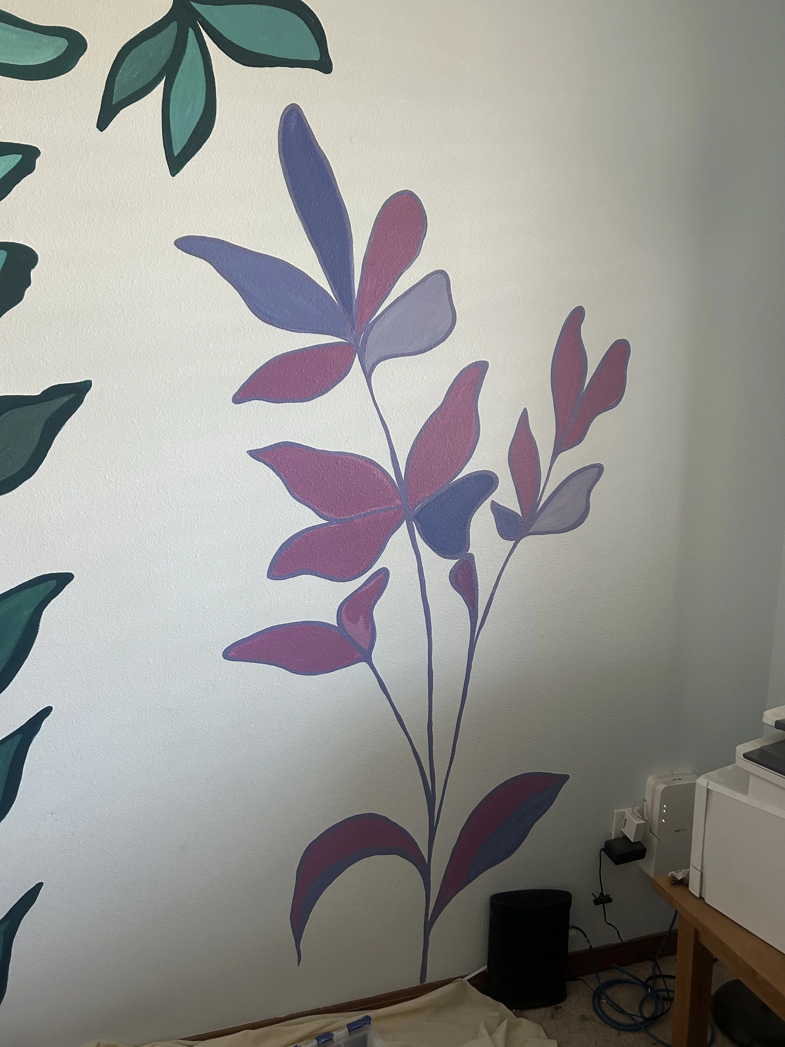 Interior wall decorated with large, colorful botanical mural featuring purple and pink leaves. There is a corner of a wooden desk with various electronic devices and cords, including a white device, a black speaker or trash bin, and power strips on the floor.