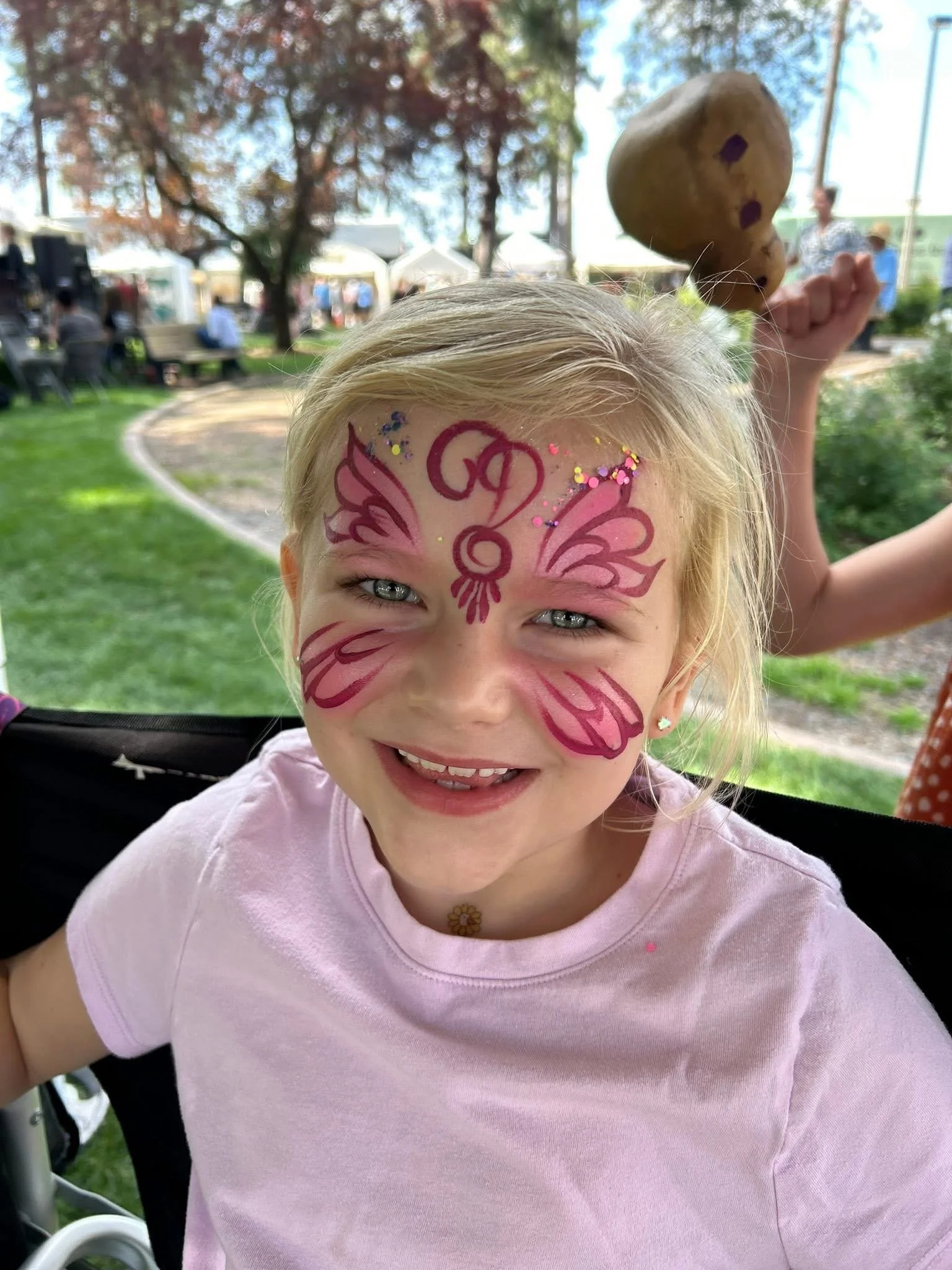 A young girl with face paint in pink and purple butterfly and floral designs on her face, smiling outdoors with a blurred park background, holding a potato with a surprised face drawn on it on her raised hand.