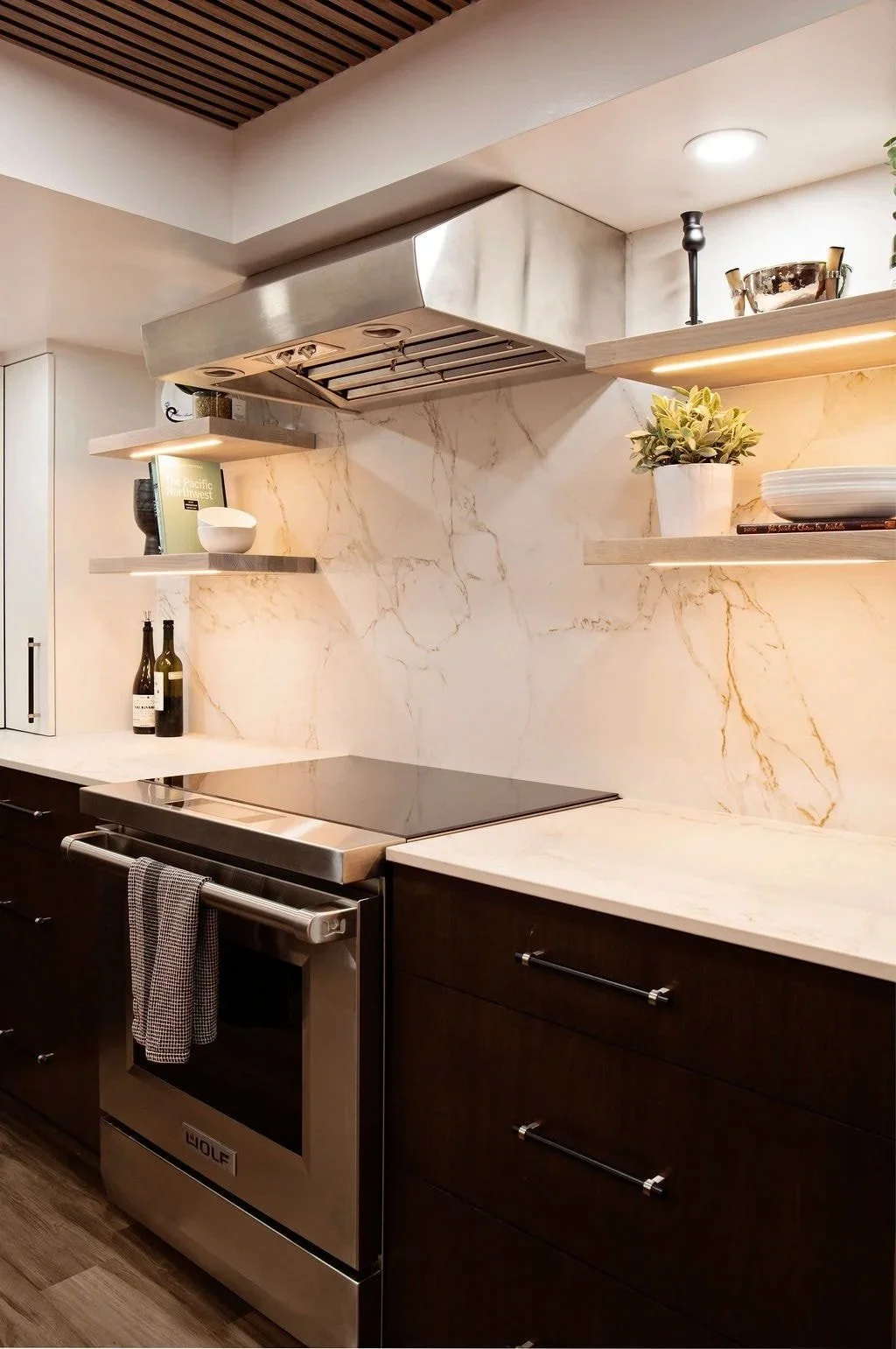 Modern kitchen with dark wood cabinets, a stainless steel Wolf oven, marble backsplash, open shelves with decorative items, a stovetop, and a ventilation hood.