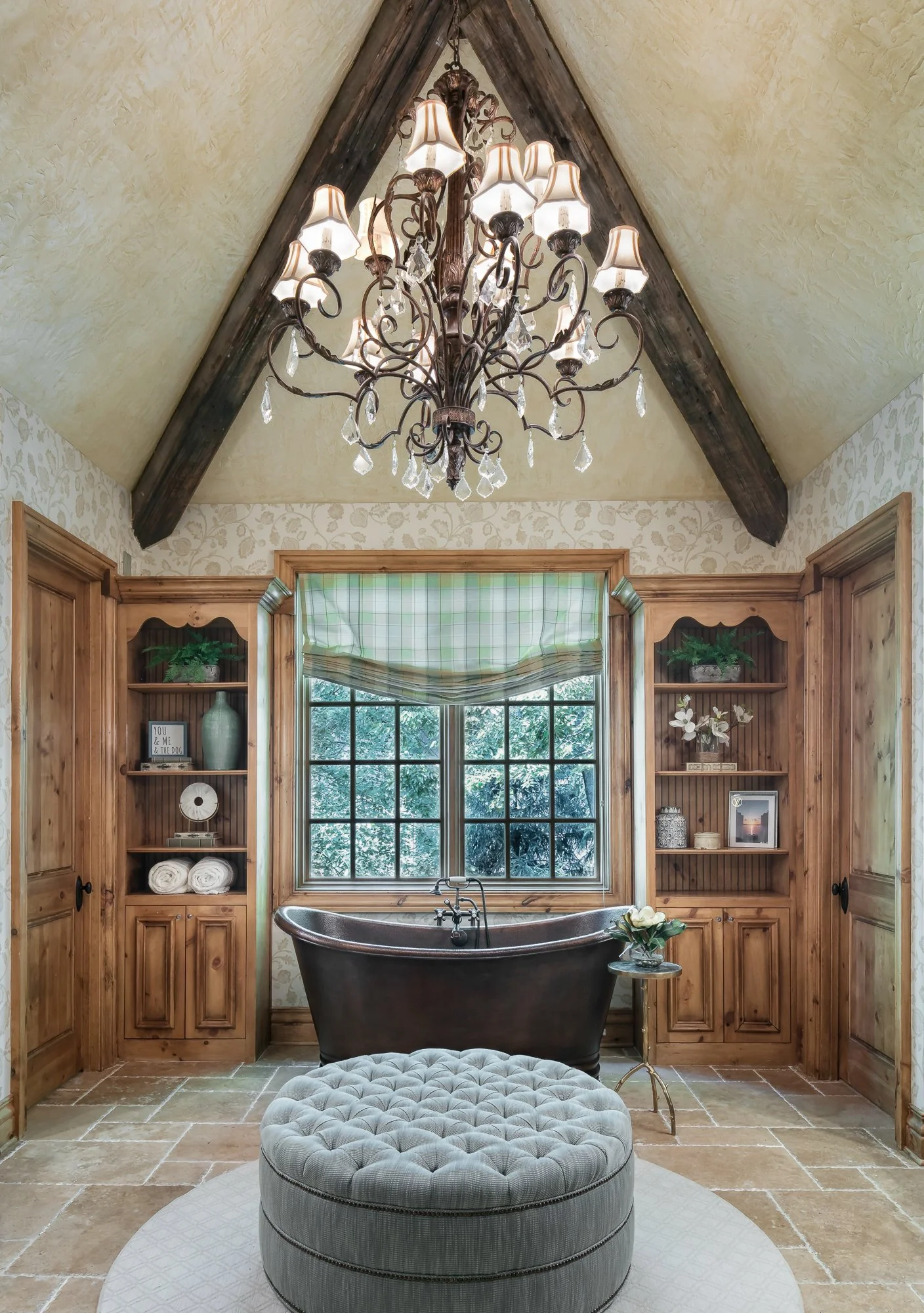 A vaulted, wood-beamed ceiling anchors this arched niche, lit by an ornate crystal chandelier. Built-in wood cabinetry lines both sides of an arched window dressed with a roman shade, and a round tufted ottoman sits centered on a patterned area rug