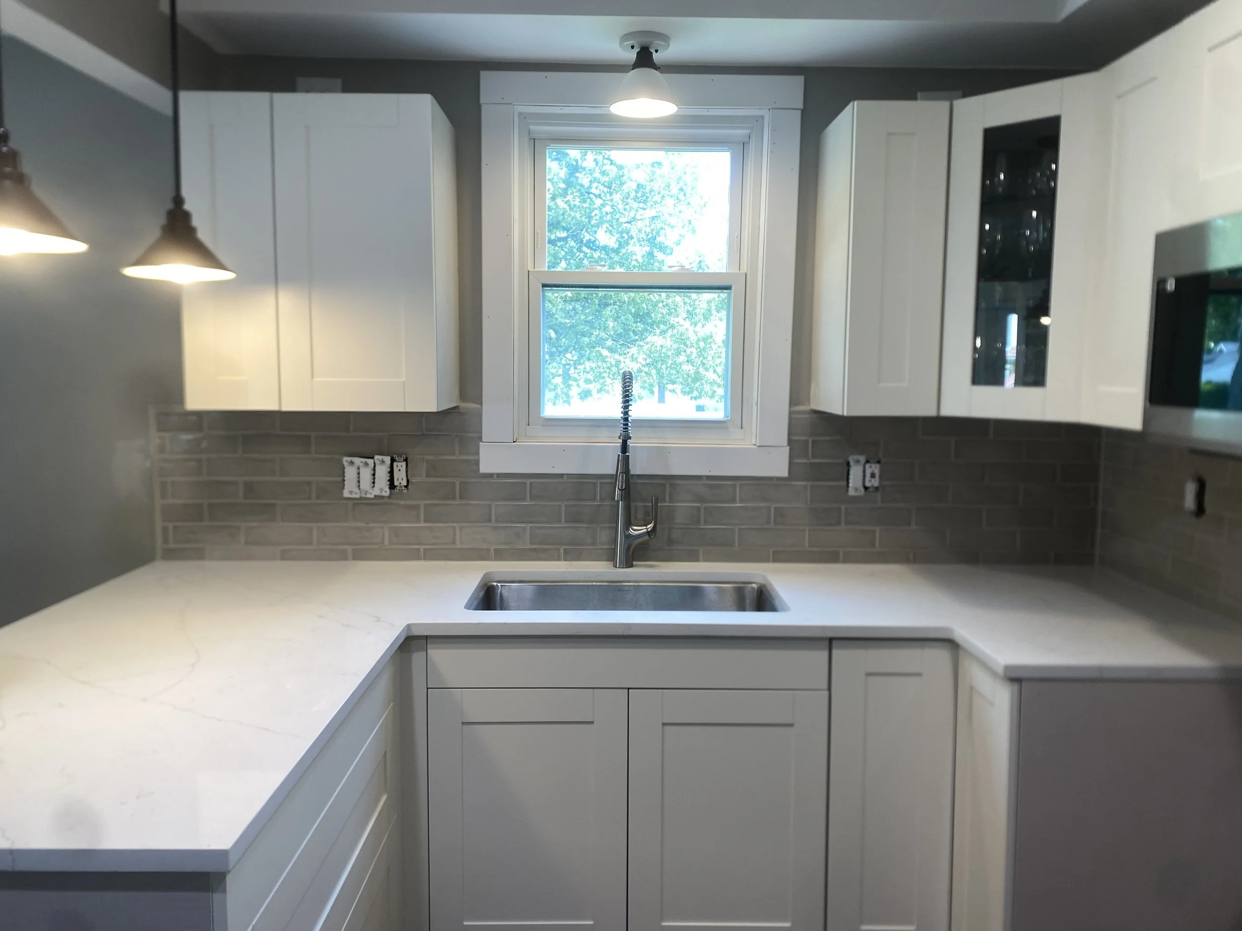 Modern kitchen with white cabinets, a large window showing green trees outside, under-cabinet lighting, a countertop with a light-colored marble pattern, and a stainless steel sink with a high-arc faucet.