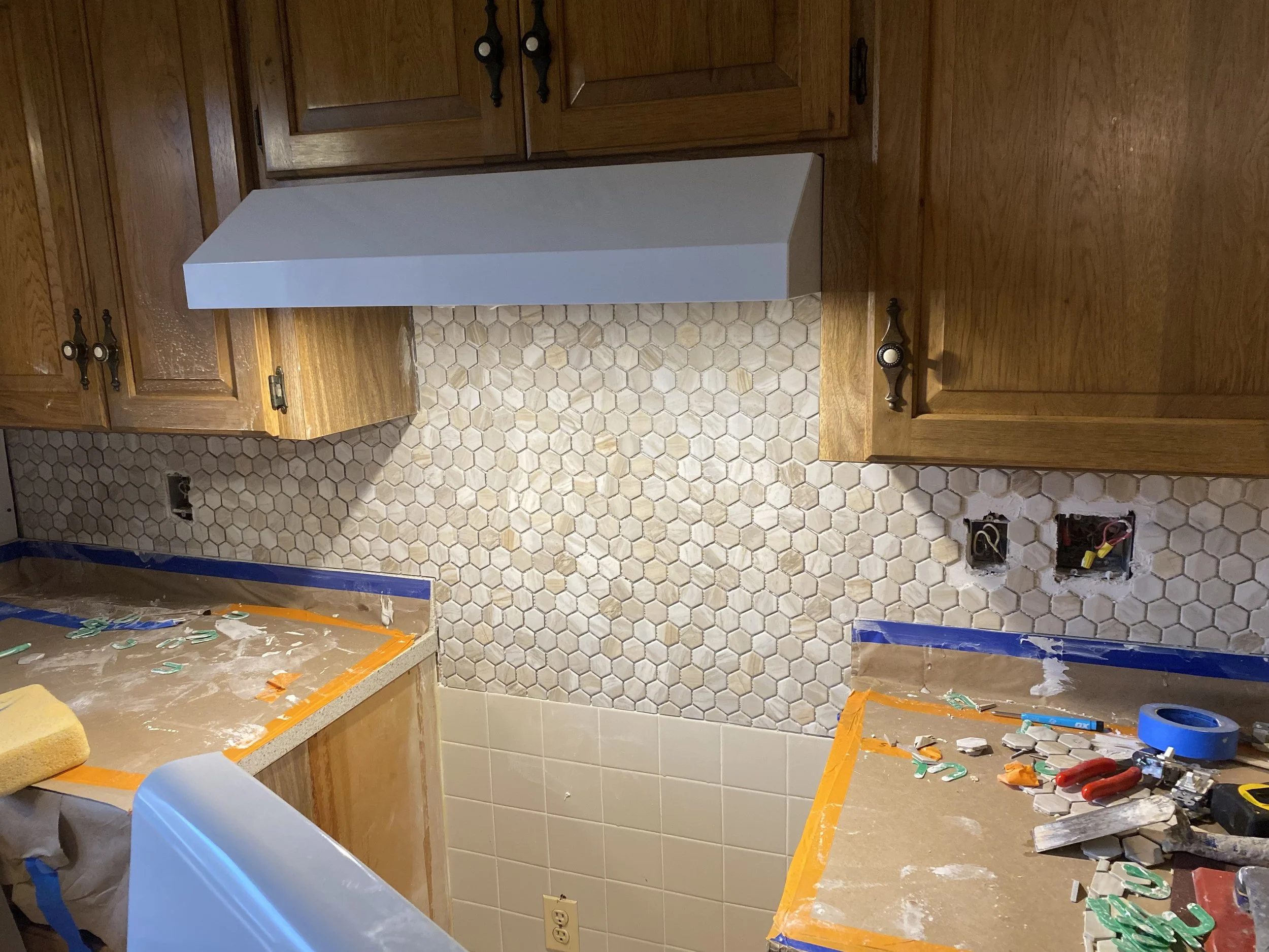 Kitchen under renovation with wooden cabinets, a hexagonal tile backsplash, and a counter covered with construction tools and materials.