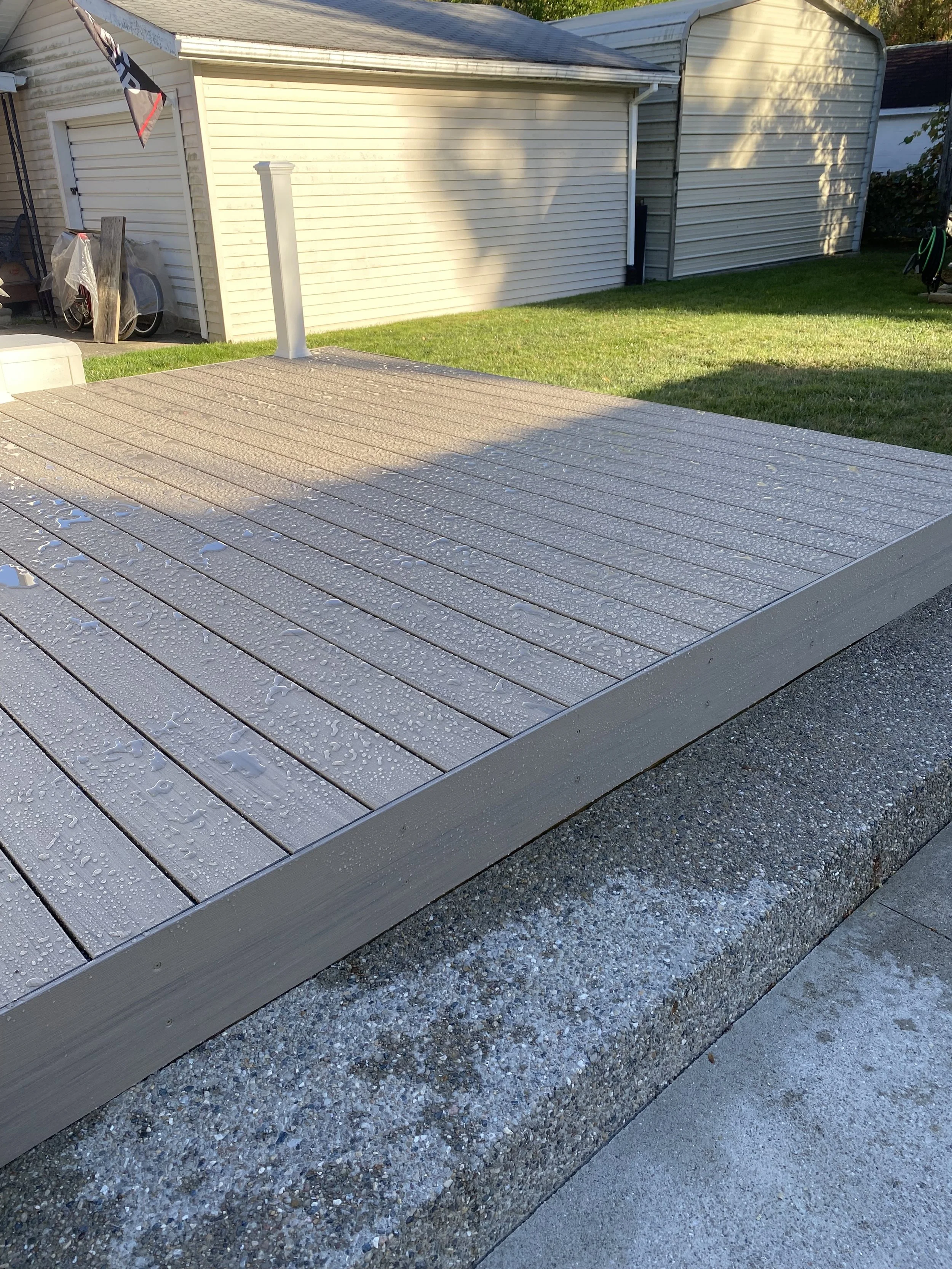 A newly installed wooden deck with water droplets, set on a concrete and gravel base, next to a grassy yard and a white shed in the background.