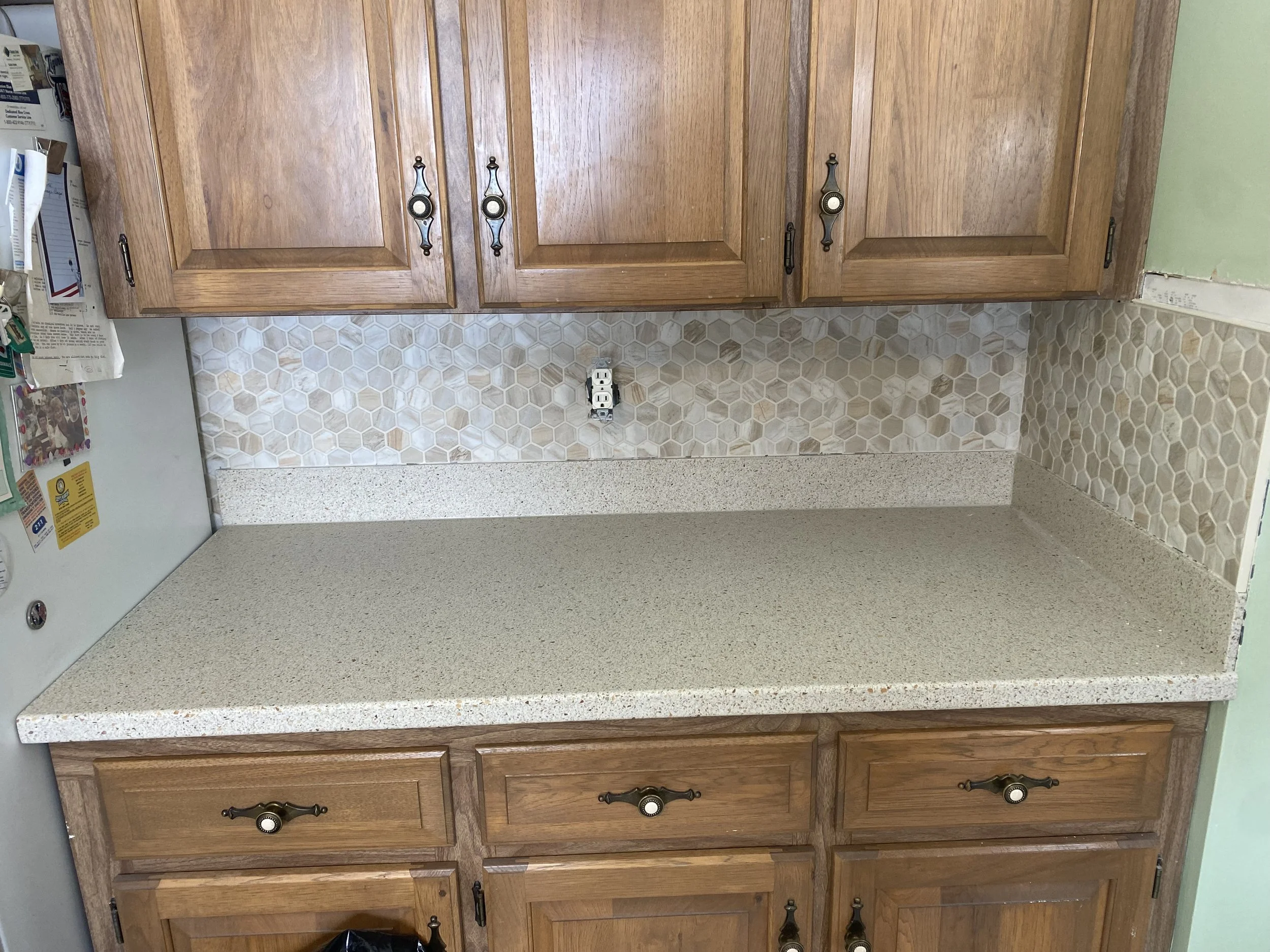 Kitchen counter with a beige speckled countertop, wooden cabinets above and below, and a two-tone hexagonal tile backsplash in beige and cream. There are miscellaneous items on the left side and an electrical outlet in the center of the backsplash.