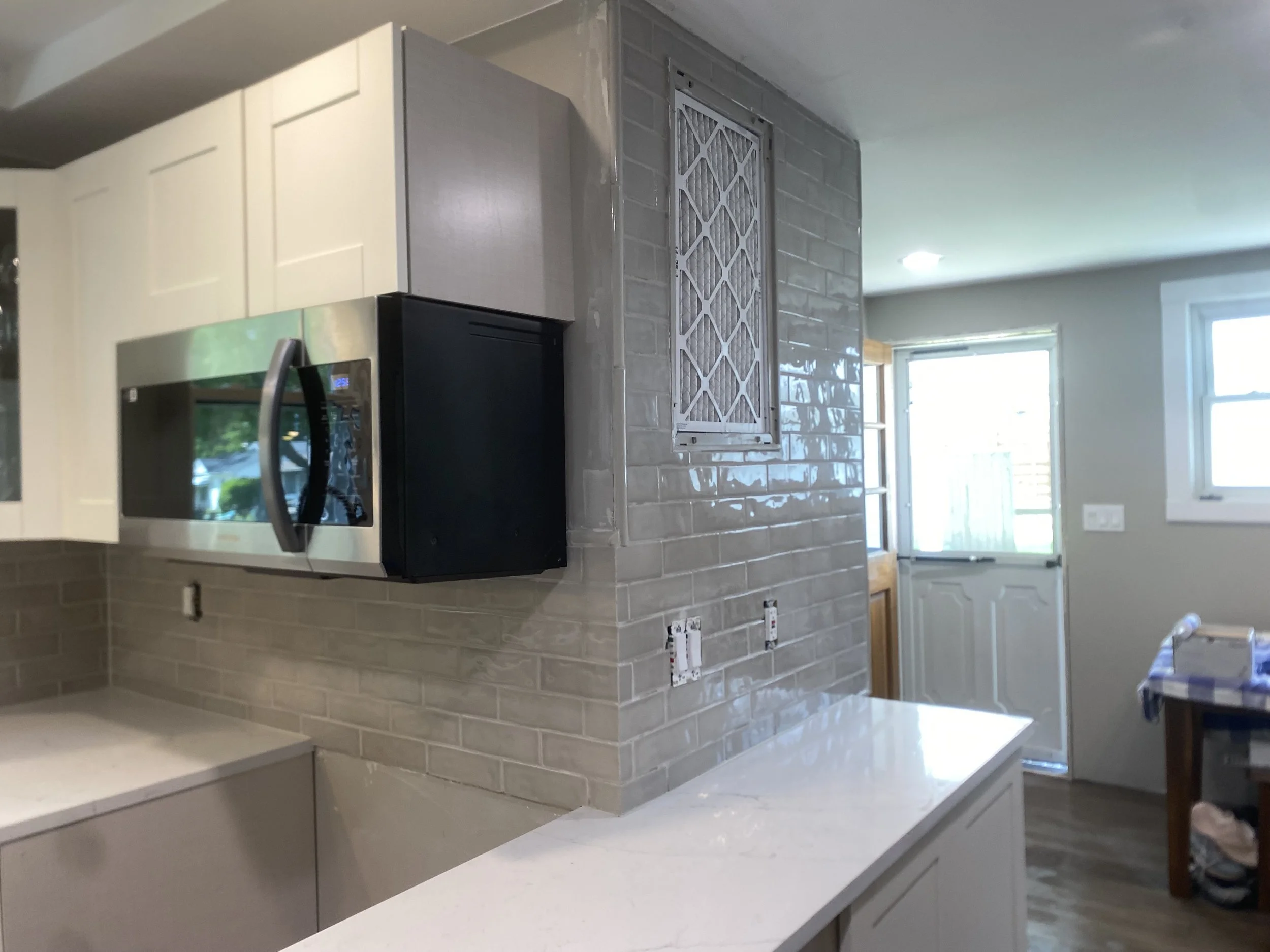 Kitchen with white cabinets, a stainless steel microwave, and a beige brick backsplash. There is a window brightening the space and electrical outlets on the wall.