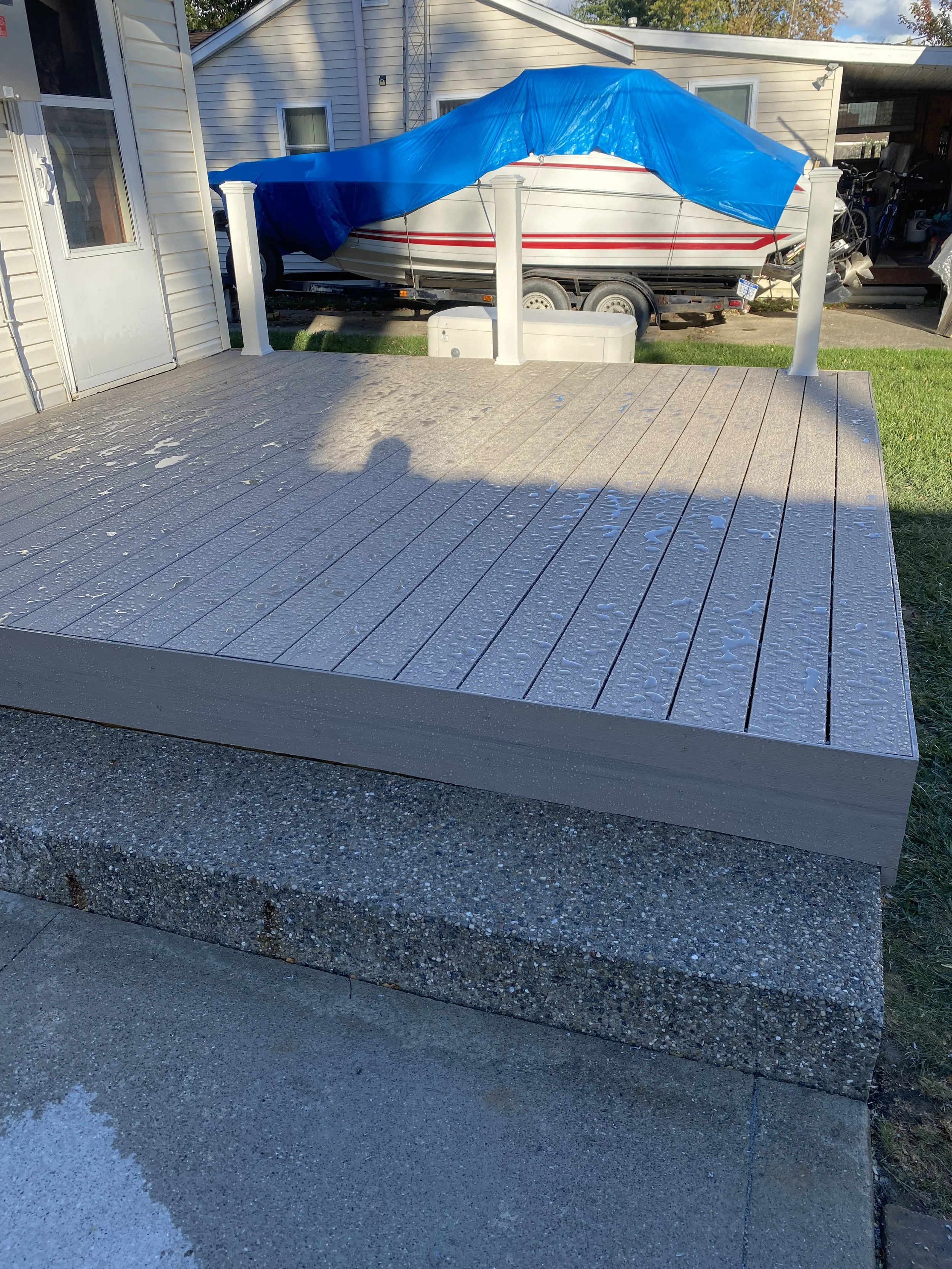 A small wooden deck with water droplets on the surface, attached to a concrete foundation. In the background, a boat covered with a blue tarp is on a trailer, next to a house with white siding.