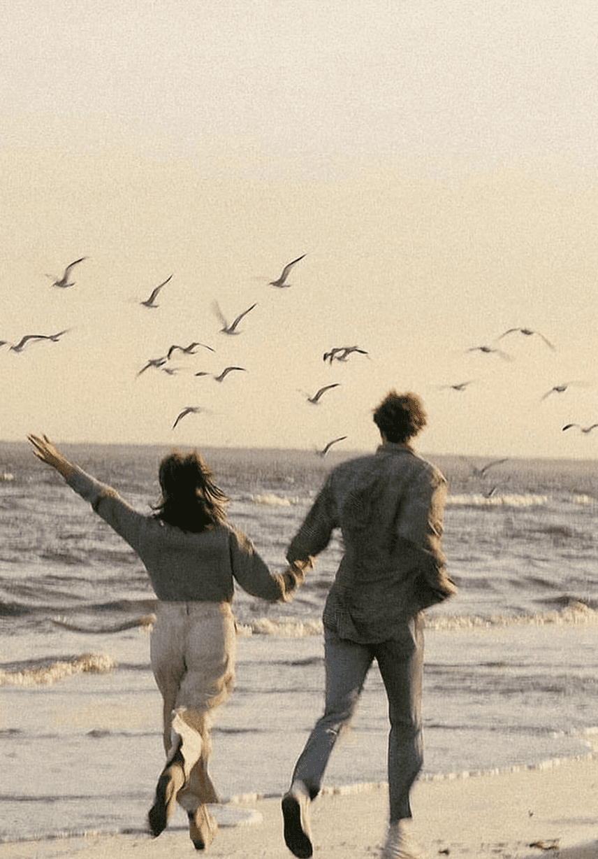 A couple holding hands and walking on the beach during sunset, with seagulls flying overhead.