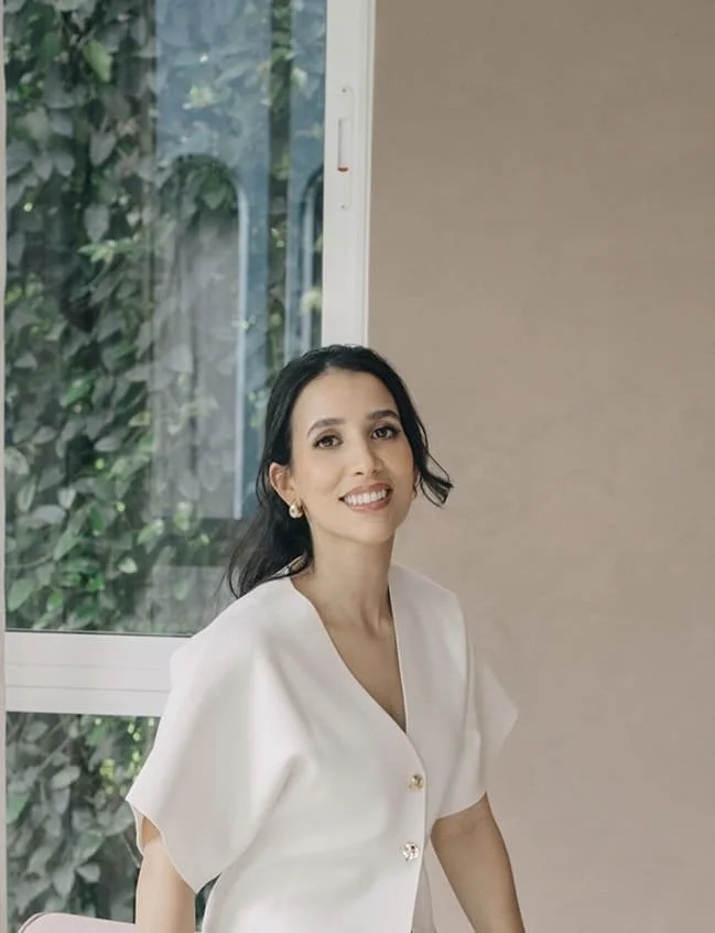 Smiling woman with long dark hair wearing a white blouse, sitting indoors near a window with green foliage outside.
