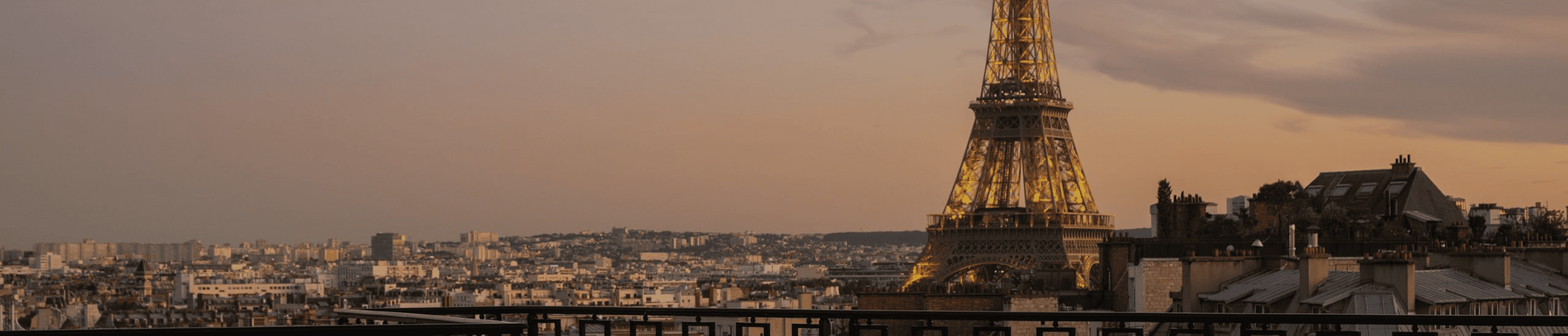 Eiffel Tower illuminated at sunset in Paris, viewed from a rooftop with surrounding buildings.