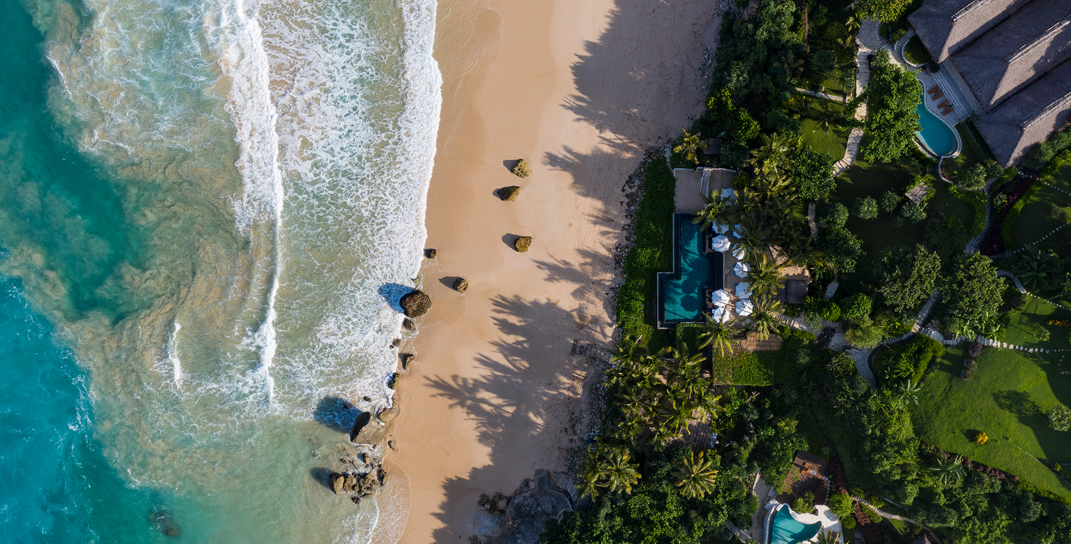 An aerial view of a tropical beach with turquoise water, white sand, rocks along the shoreline, and a luxurious resort with pools and lush green trees.