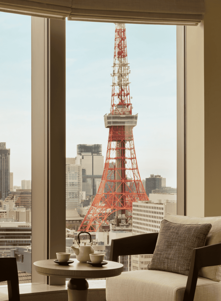 Interior of a room with a small round table set with a teapot and two cups, next to a chair with a cushion, overlooking the Tokyo Tower through large windows.