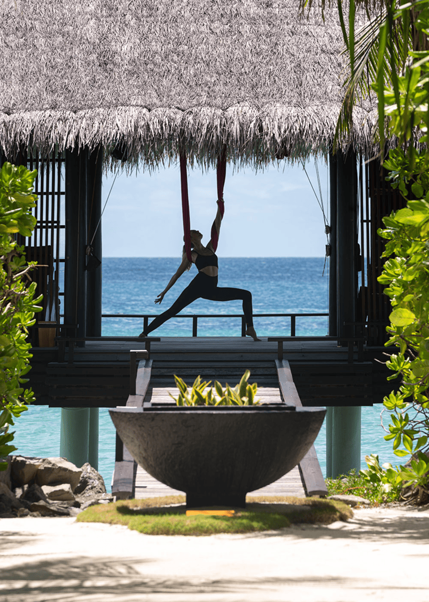 A woman practicing aerial yoga on fabric under a thatched roof on a beach with ocean in the background.