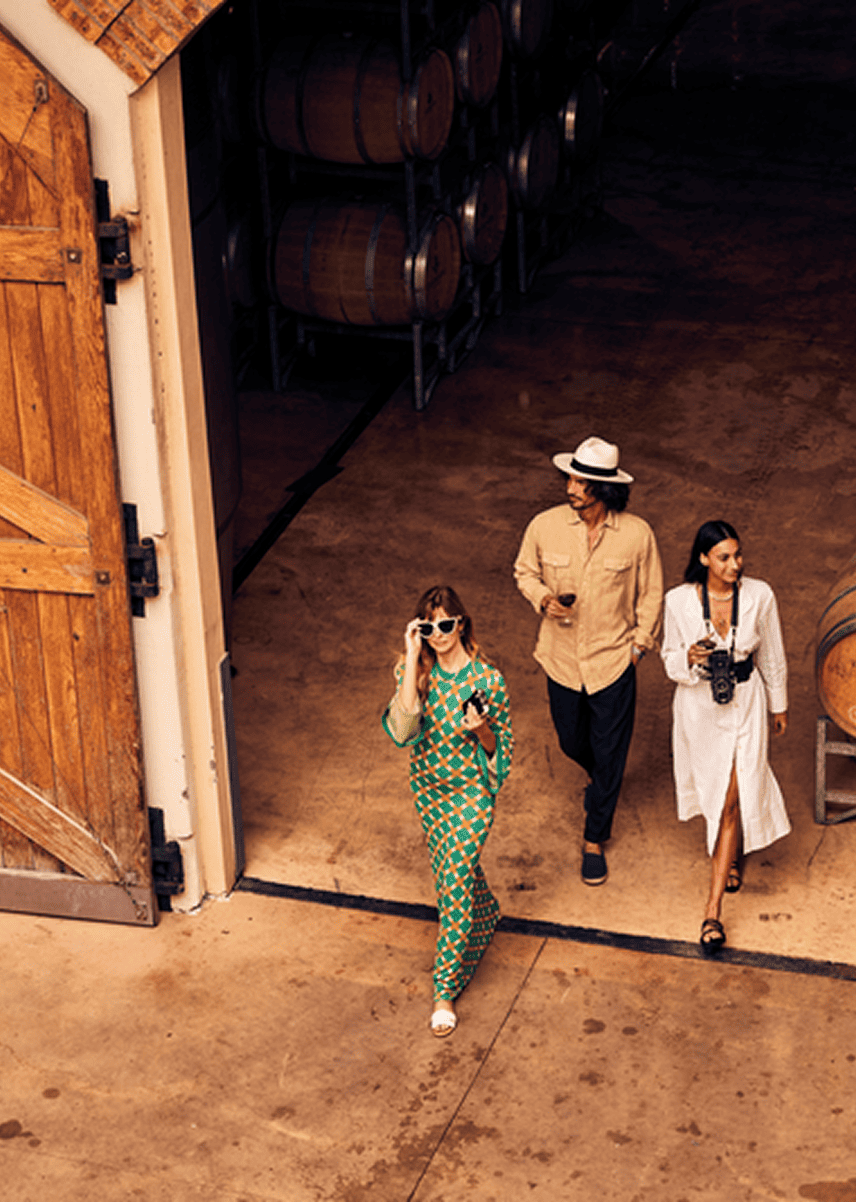 Three women walking inside a winery, one wearing sunglasses and a colorful jumpsuit, another in a beige shirt and hat, and the third with a camera around her neck.