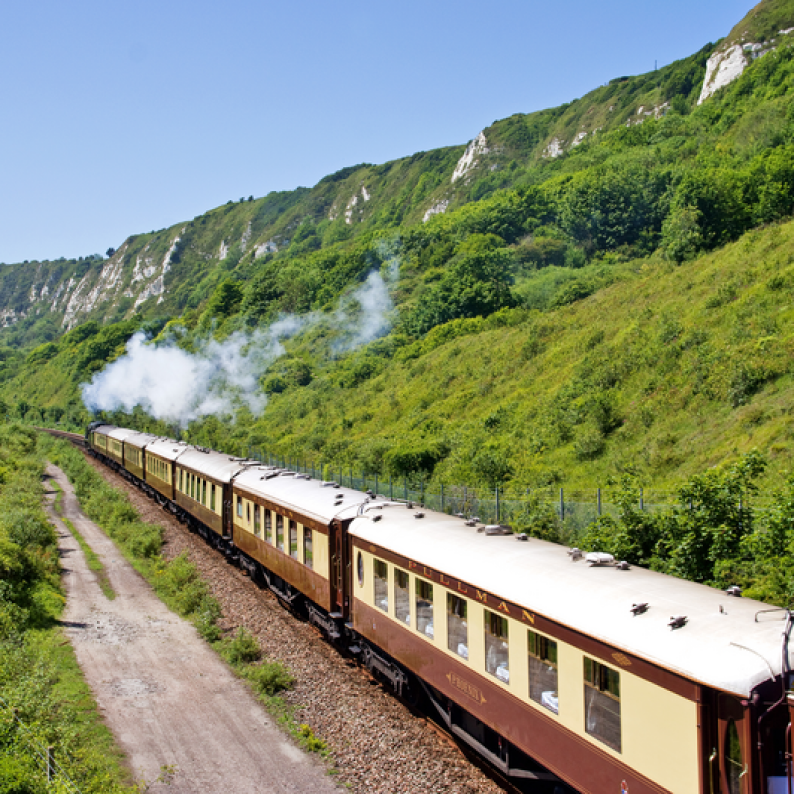 A vintage passenger train traveling along a scenic mountain track, surrounded by lush green hills and cliffs under a clear blue sky.