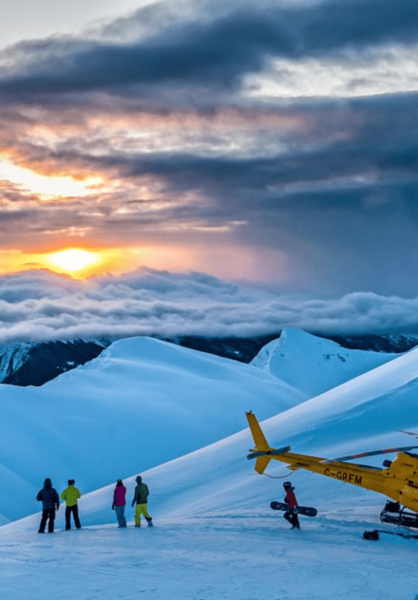 A group of four people standing on snow-covered ground with a yellow helicopter nearby, in a cold mountainous landscape during sunset with clouds and snow-capped peaks in the background.