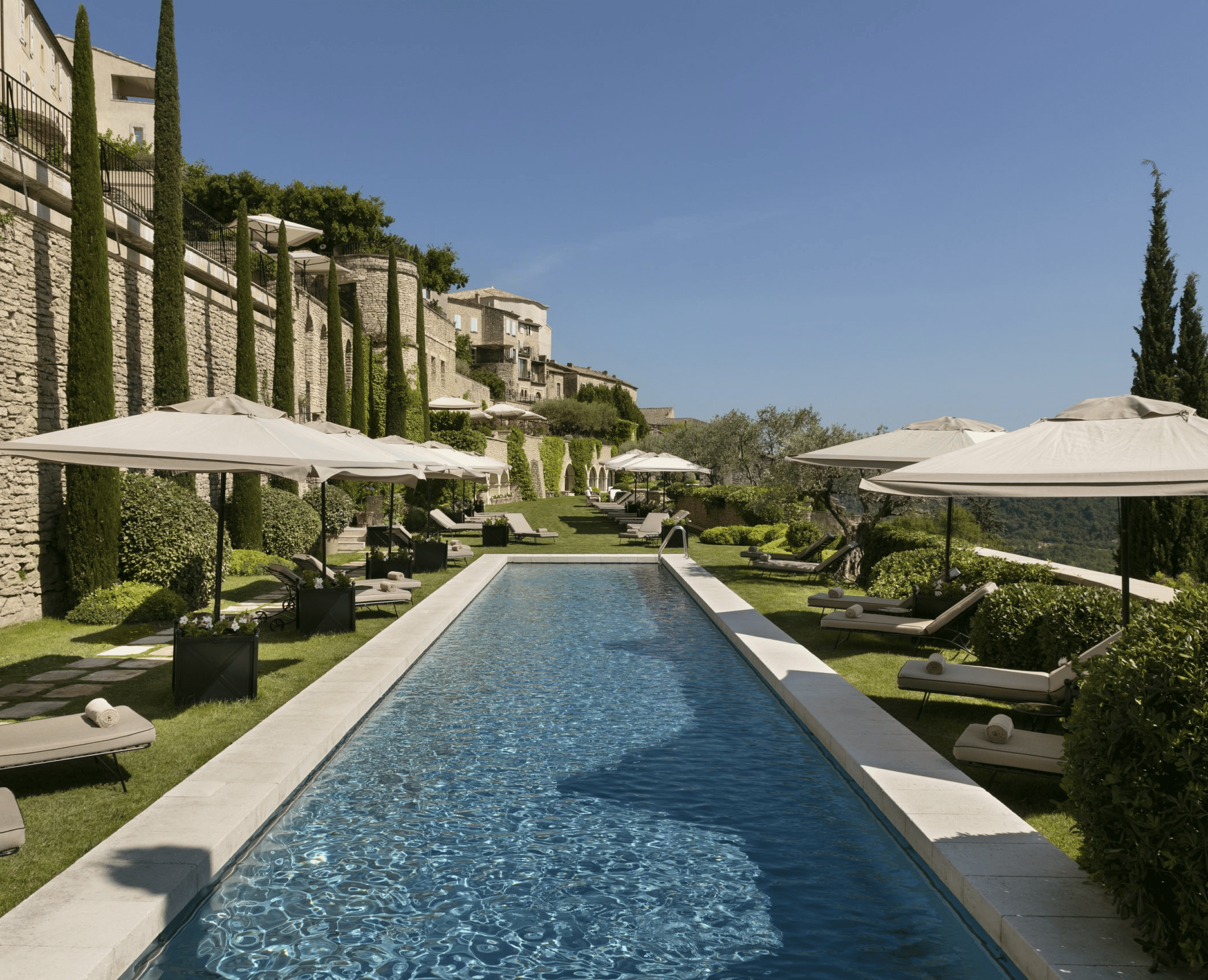 A luxurious outdoor swimming pool surrounded by lounge chairs with rolled towels, large white umbrellas, and lush greenery in a hillside setting with stone buildings, tall cypress trees, and a clear blue sky.