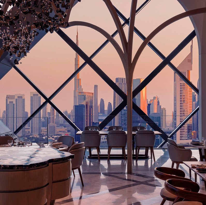 Interior of a modern high-rise restaurant with large panoramic windows showing an urban city skyline with skyscrapers at sunset.