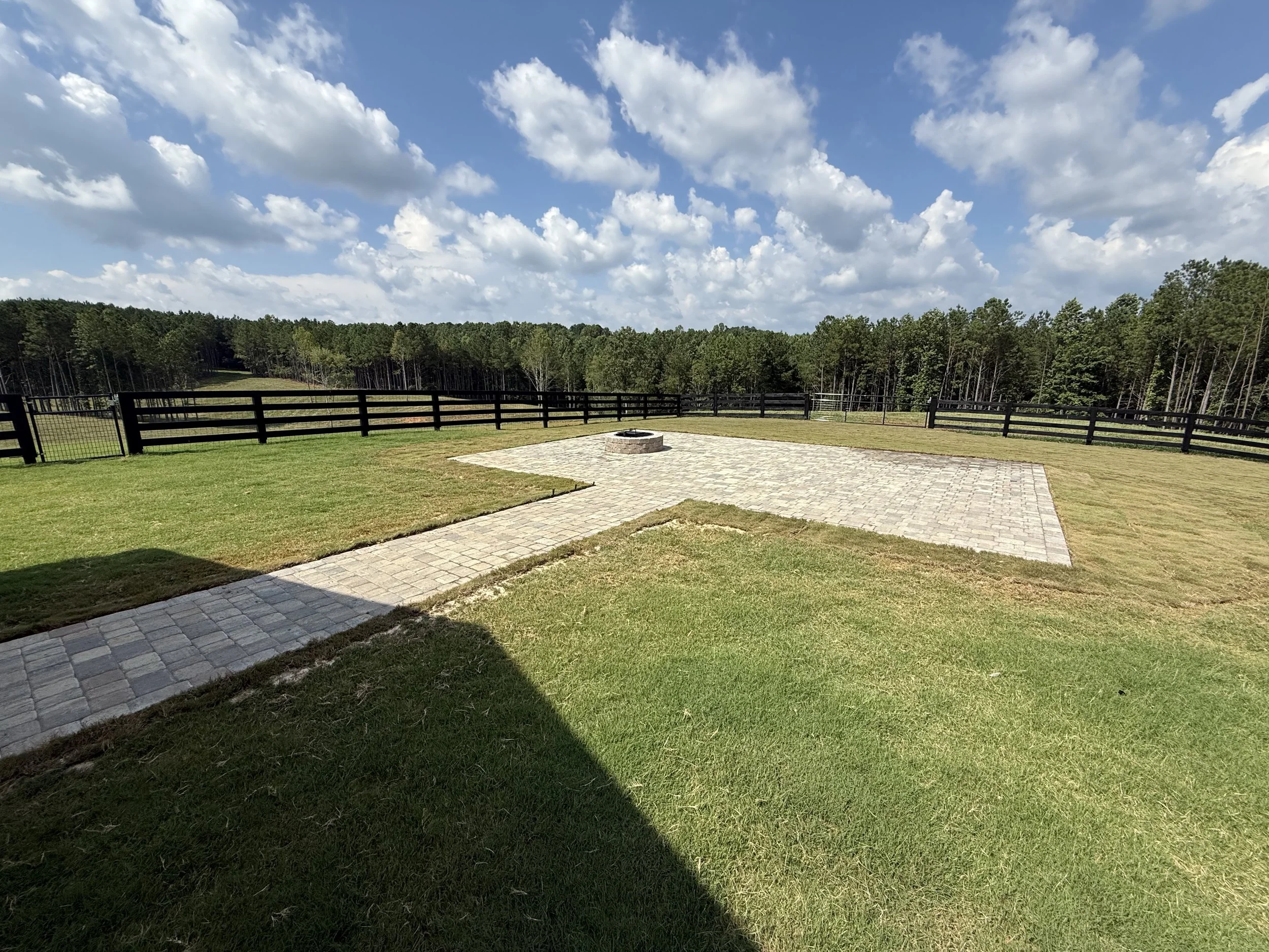 A backyard with a grassy lawn, a paved patio area with a fire pit, black fencing, and trees in the background under a partly cloudy sky.