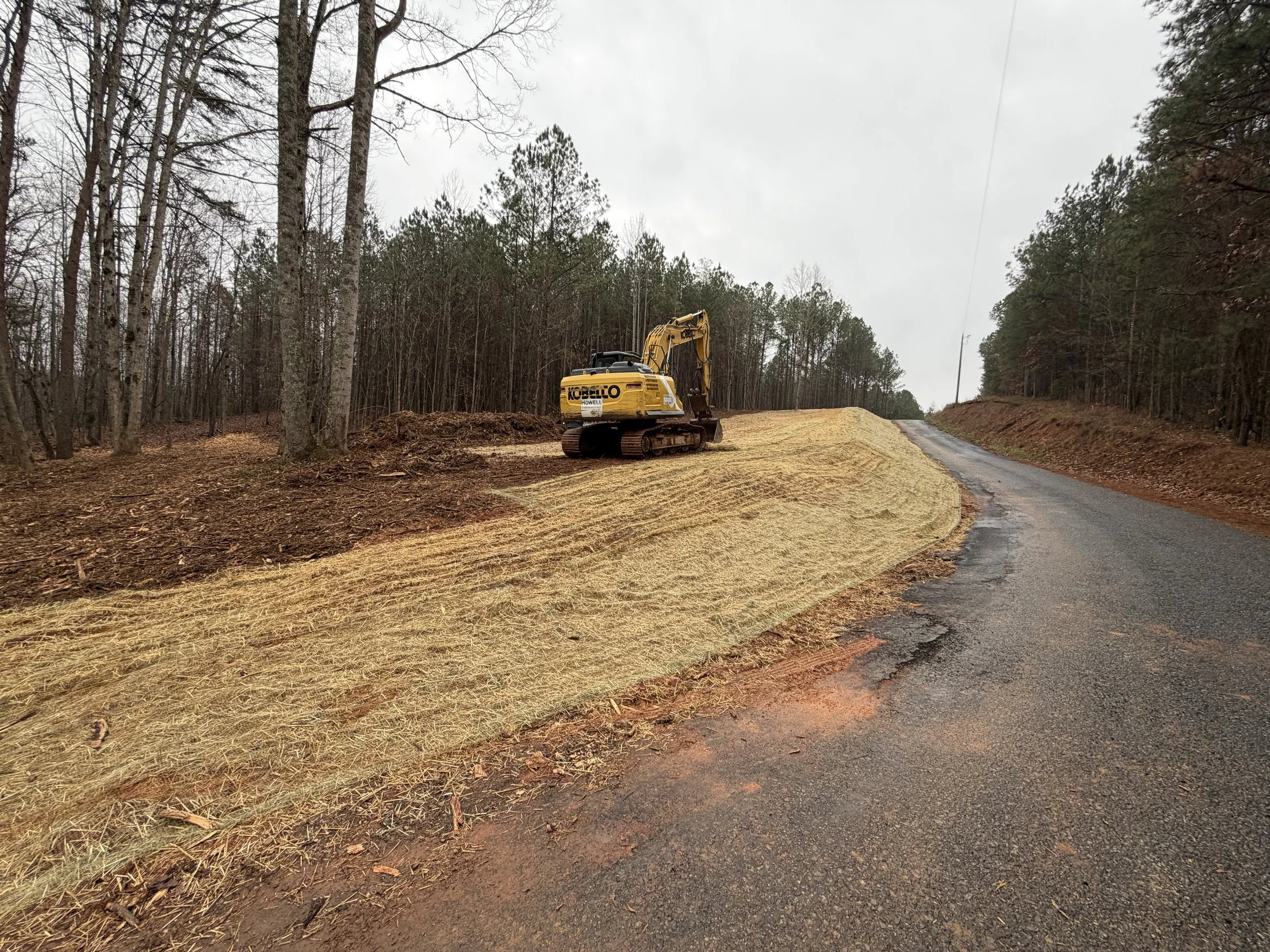 A yellow excavator parked on a grassy cleared area beside a paved rural road, with trees in the background on overcast day.