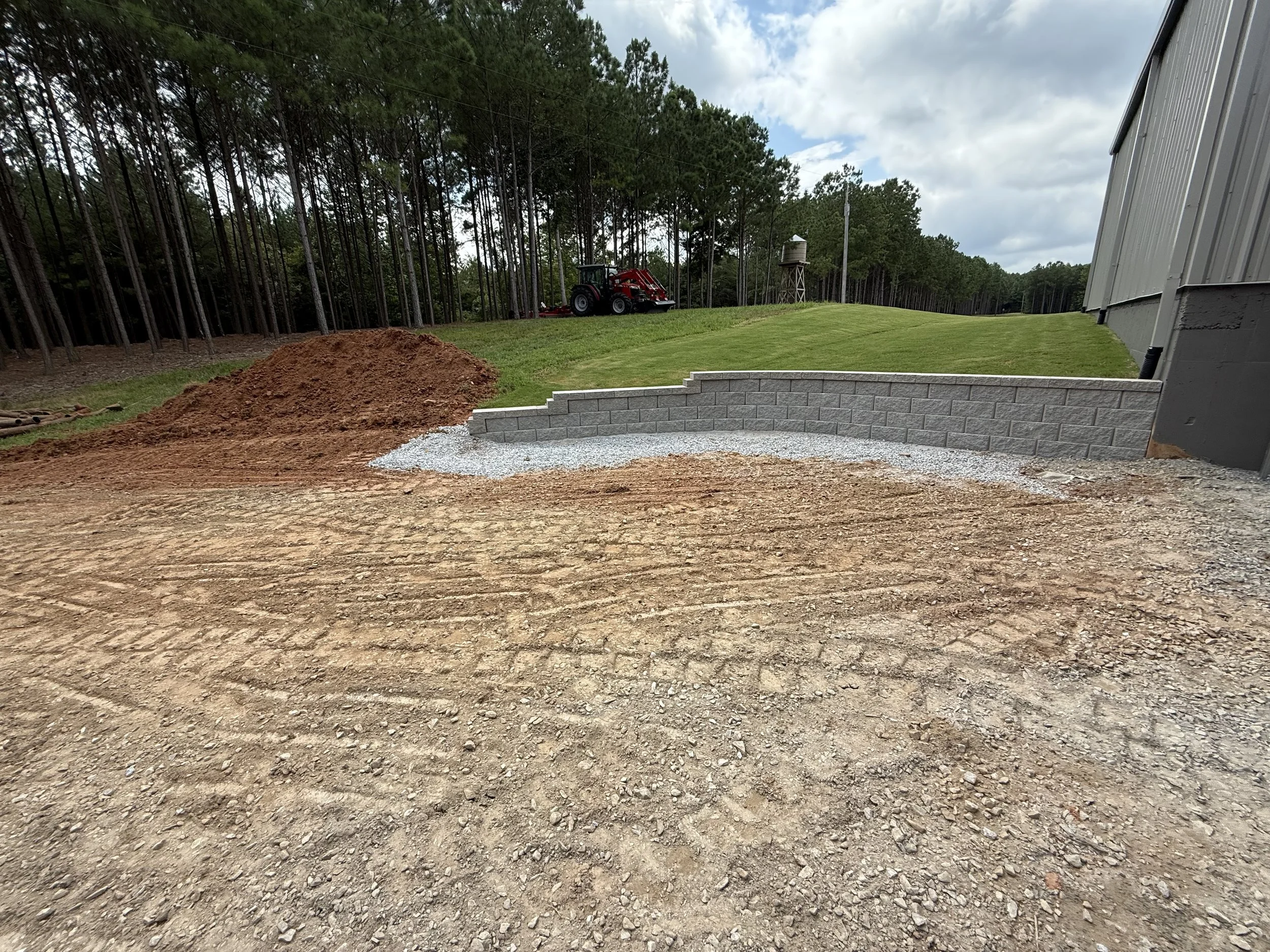 A construction site with a gravel pathway, a partially built brick wall, a mound of dirt, and a tractor in the distance near a wooded area and open grassy field.