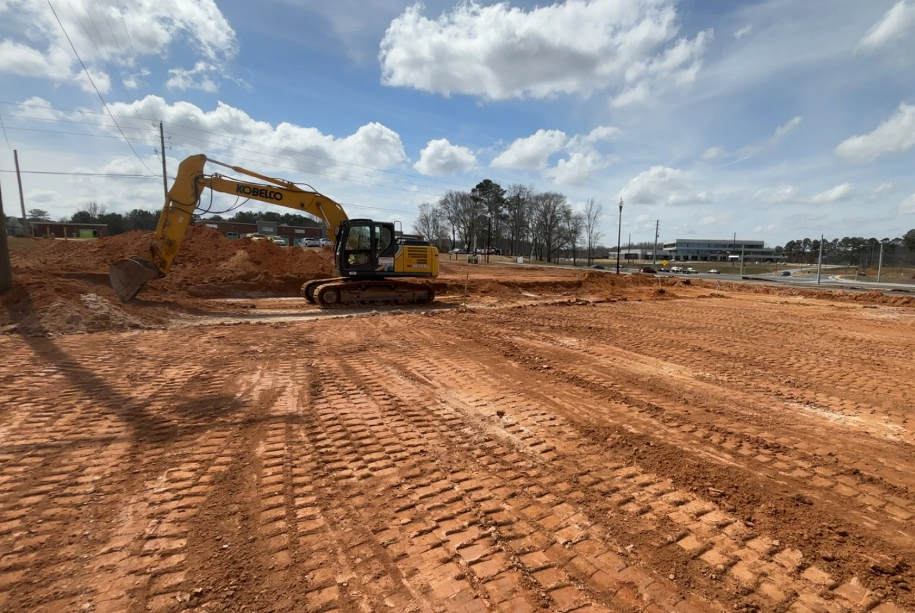 Construction site with a yellow excavator moving dirt on a cleared area under partly cloudy skies. There are roads and buildings in the background.