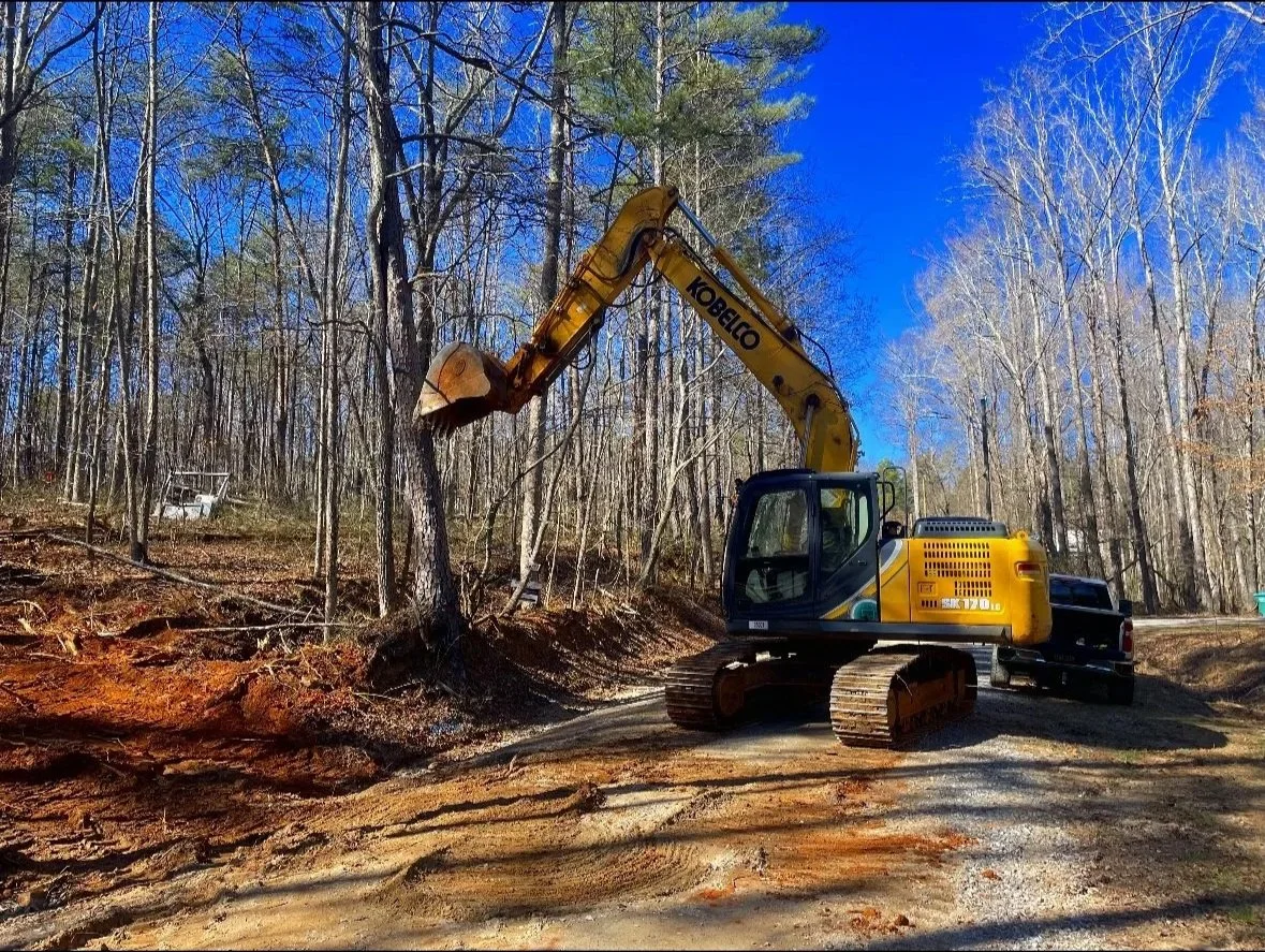 A yellow excavator with tracks is demolishing a tree in a forested area with leafless trees, blue sky, and dirt ground.